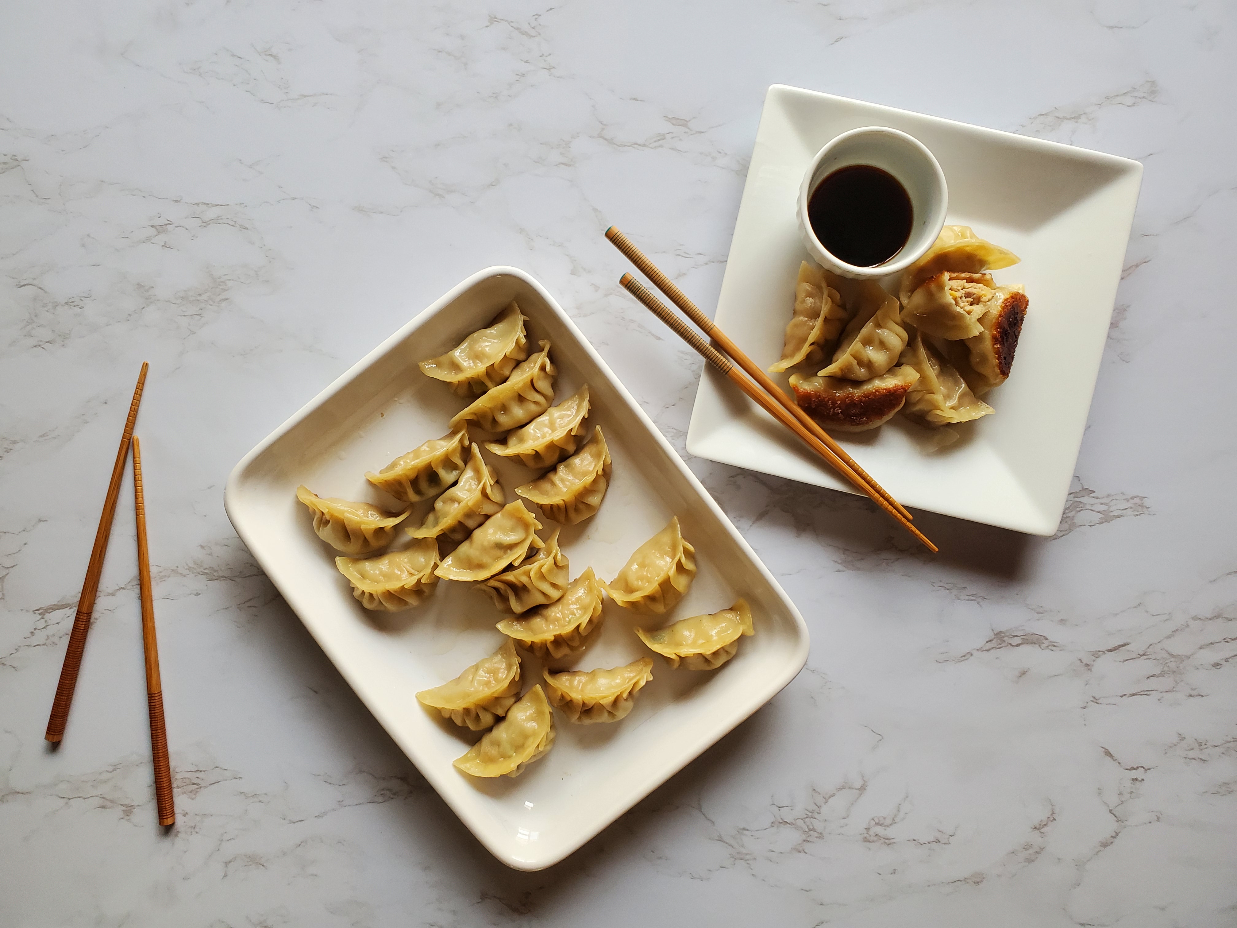 A large rectangular plater of Chinese Potstickers sits next to a square plate of potstickers with a small bowl of dark brown dumpling sauce. A pair of chopsticks rest on the square plate and on the left of the platter on the white marble coutertop.