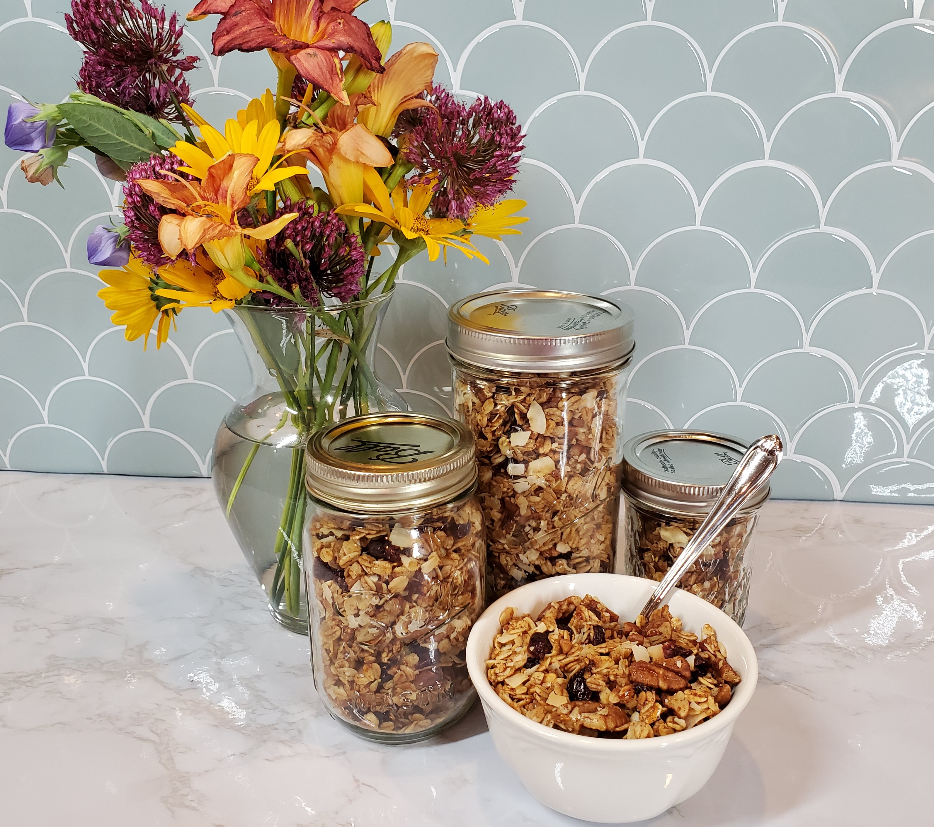 A bowl of pecan/craisin granola in a white bowl with a spoon sits in front of three Mason jars of granola on a white granite counter top. Behind it is a vase filled with yellow, pink and purple flowers and a shell turquoise tile backsplash.