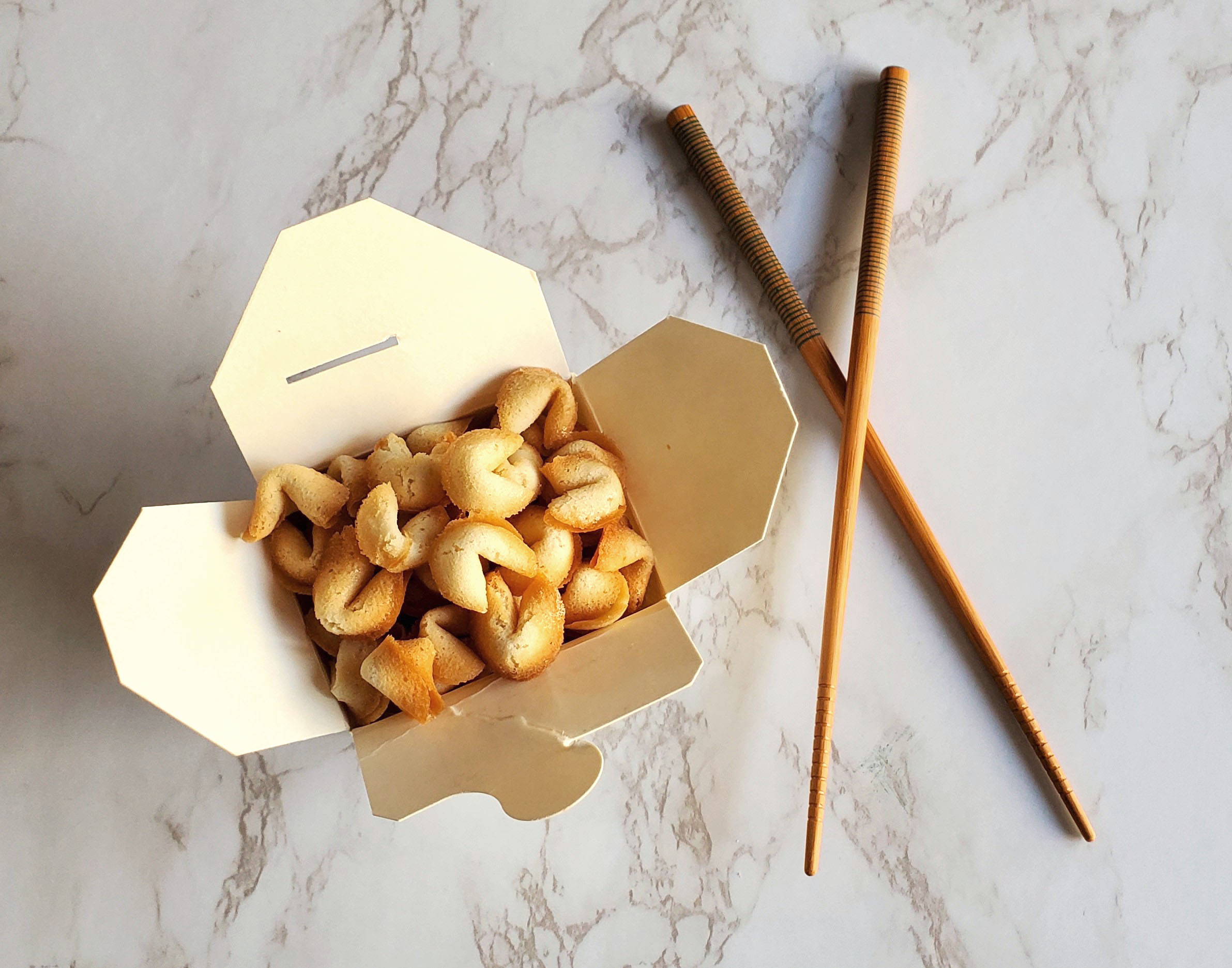 There is a small white Chinese takeout box filled with Fortune Cookie Cereal, overflowing out of it. To the right there is a pair of chopsticks with a green lined pattern at the top. The chopsticks are criss-crossed on top of the white marble countertop.