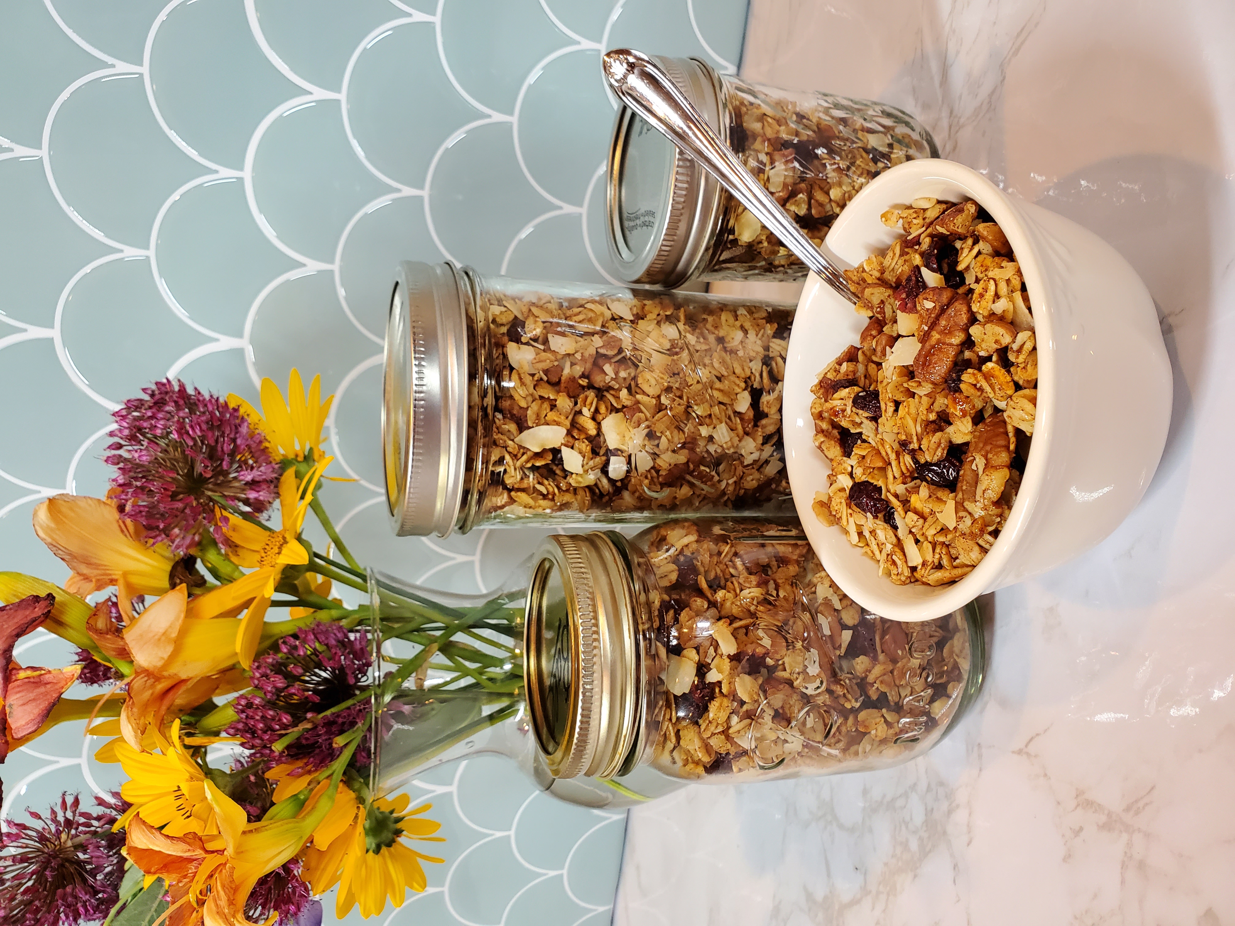 Three jars of granola sit behind a bowl of granola. Bits of pecans and craisins are visible. There is a siler spoon sticking out of the bowl and there is a vase full of flowers behind the scene, yellow, pink and purple.