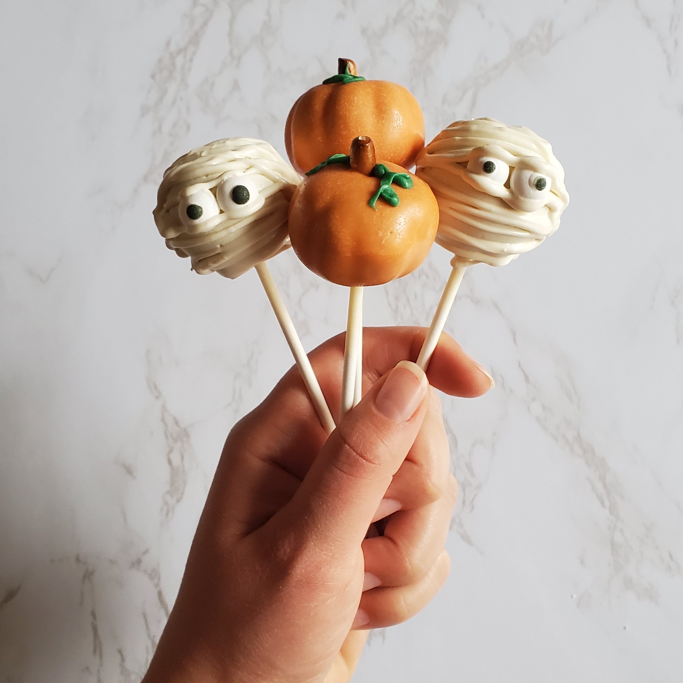 A hand up against a white marble background holds up four cake pops, two have been decorated like pumpkins and the other two have been decorated as mummies using chocolate.
