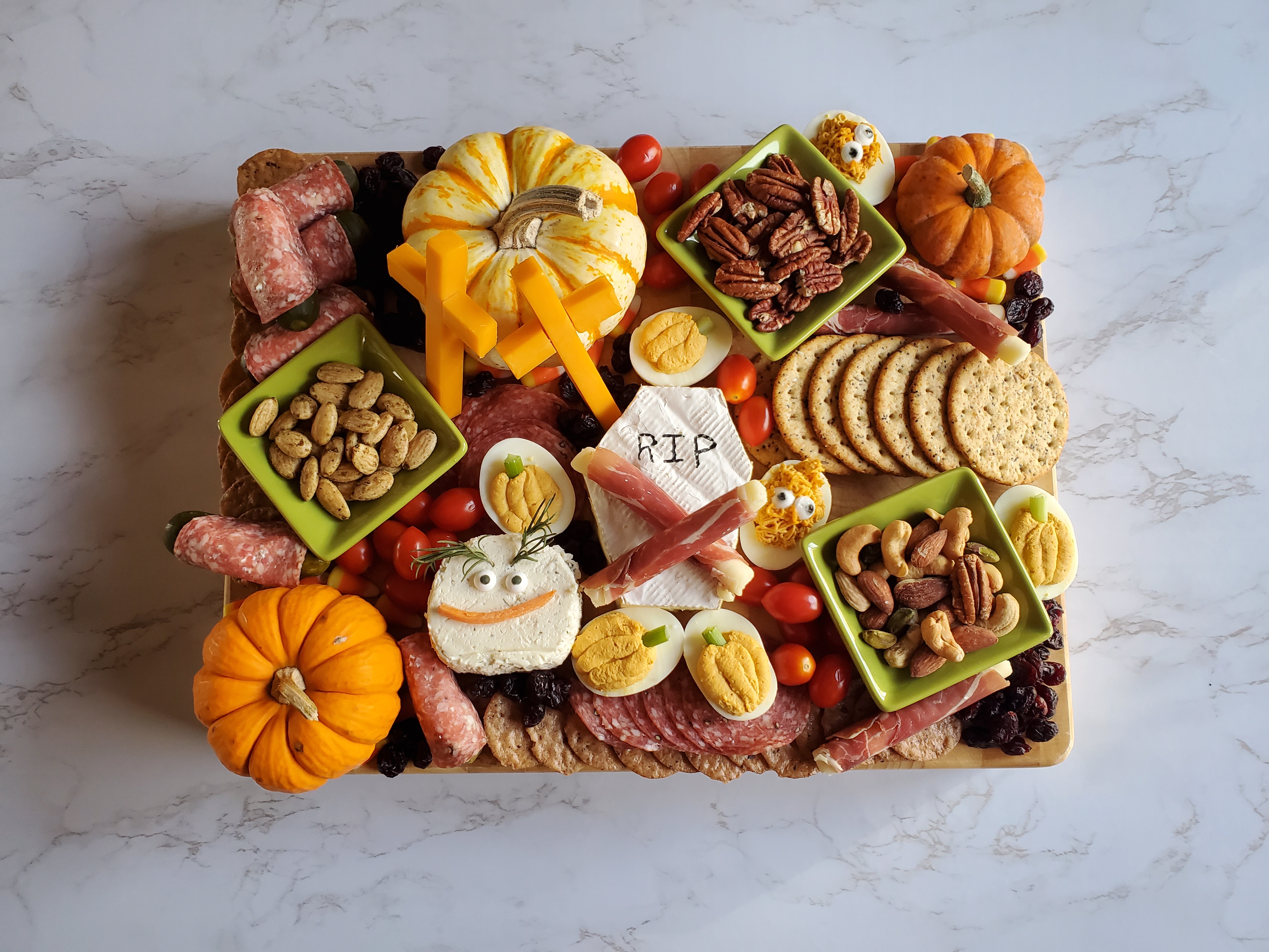 A large charcuterie board filled with a brie tombstone, mini pumpkins, cheese stick crosses, pumpkin deviled eggs, nut, dried meats and grape tomatoes on a marble countertop.