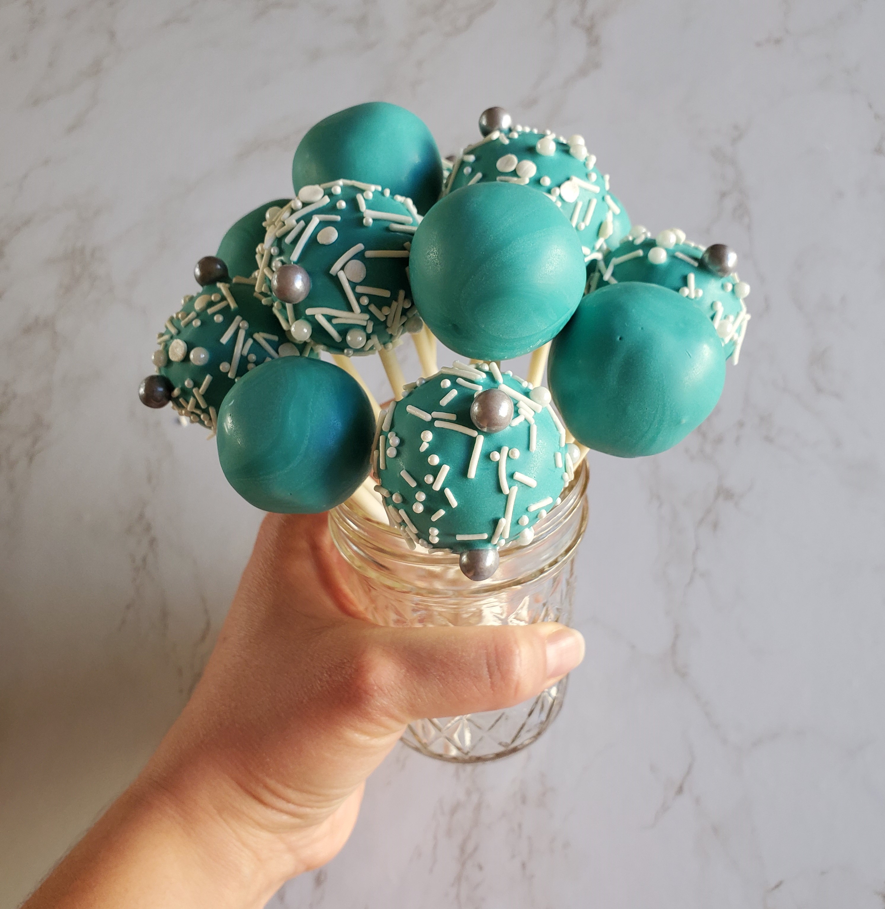 A bouquet of turquoise sprinkled cake pops in a glass jar, held up by a hand in front of a marble background.