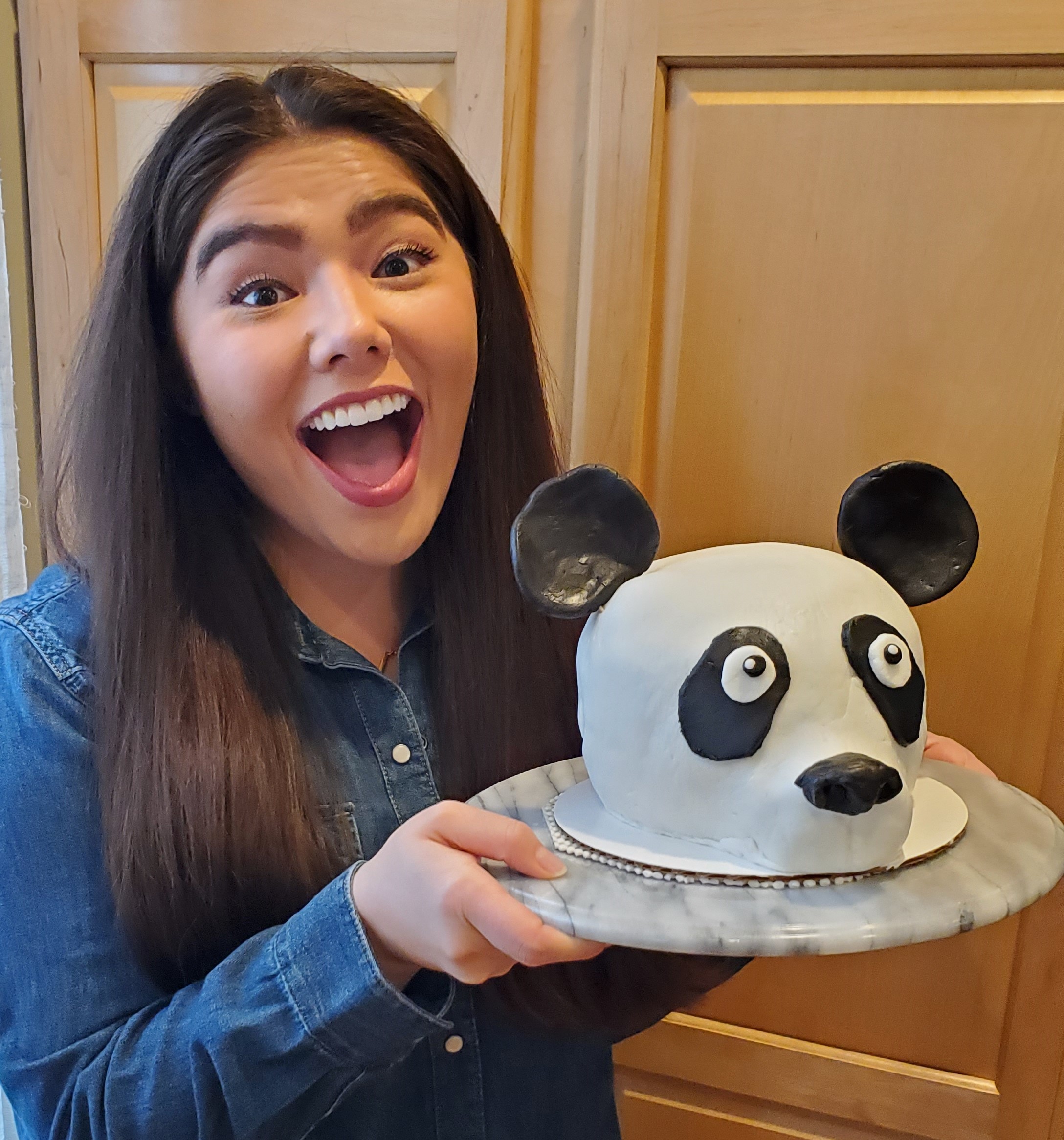 Me holding the panda cake with a "wow" expression. Behind me is a wooden cupboard.