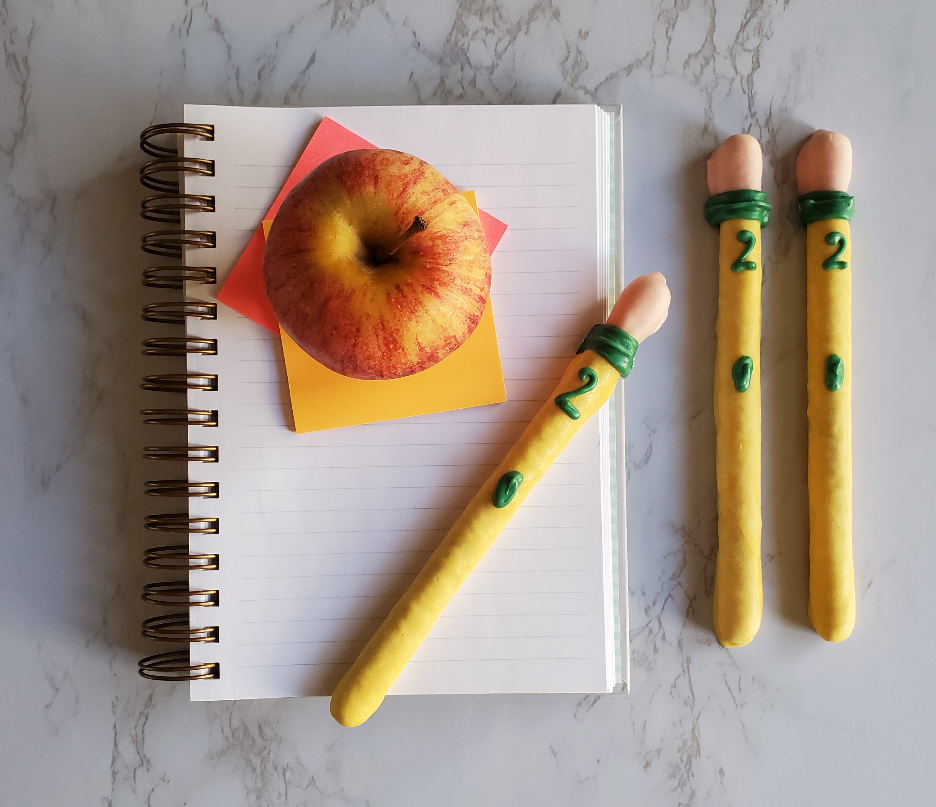 Candy "pencils" made from pretzel rods lay on top of a school ruled notebook and post-its with a lunch room style apple on top of a marble counter.