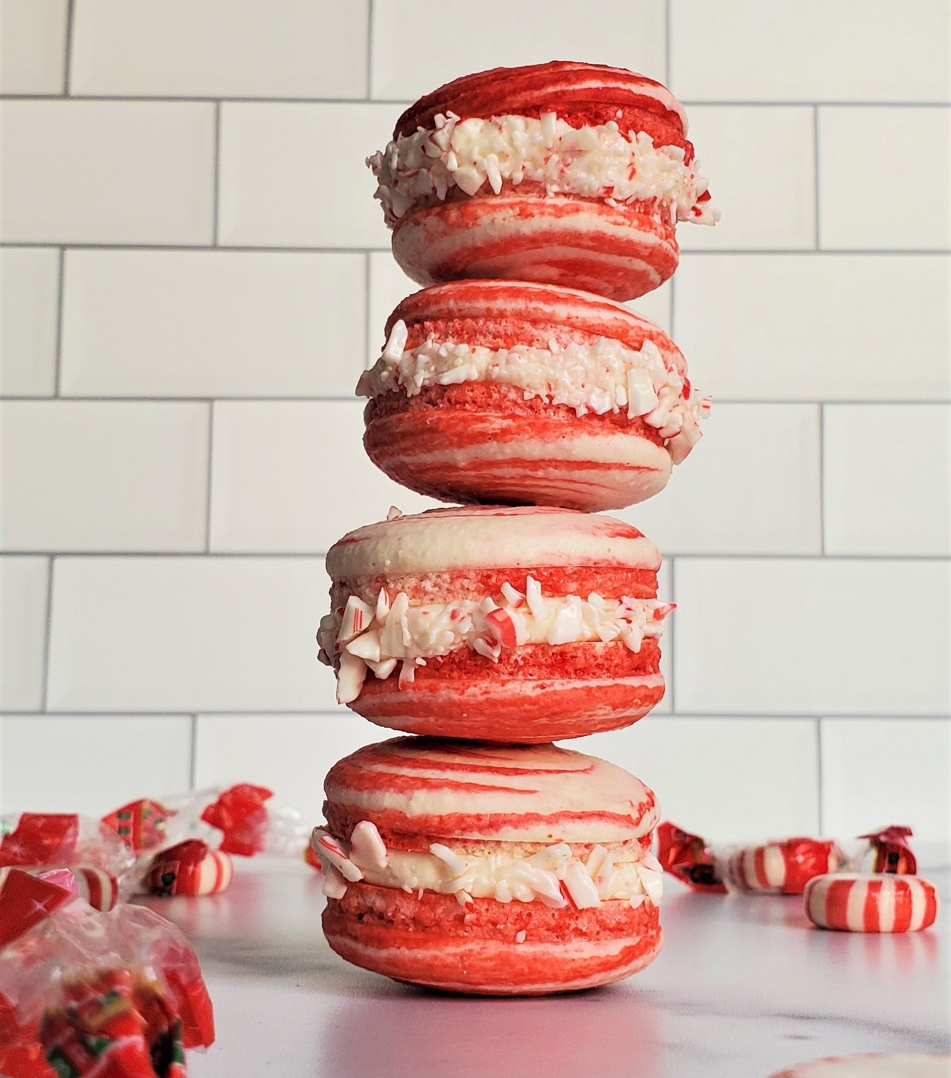 A stack of 4 peppermint macarons, up against a subway tile background, on a white marble surface. Surrounded by peppermints and their wrappers.