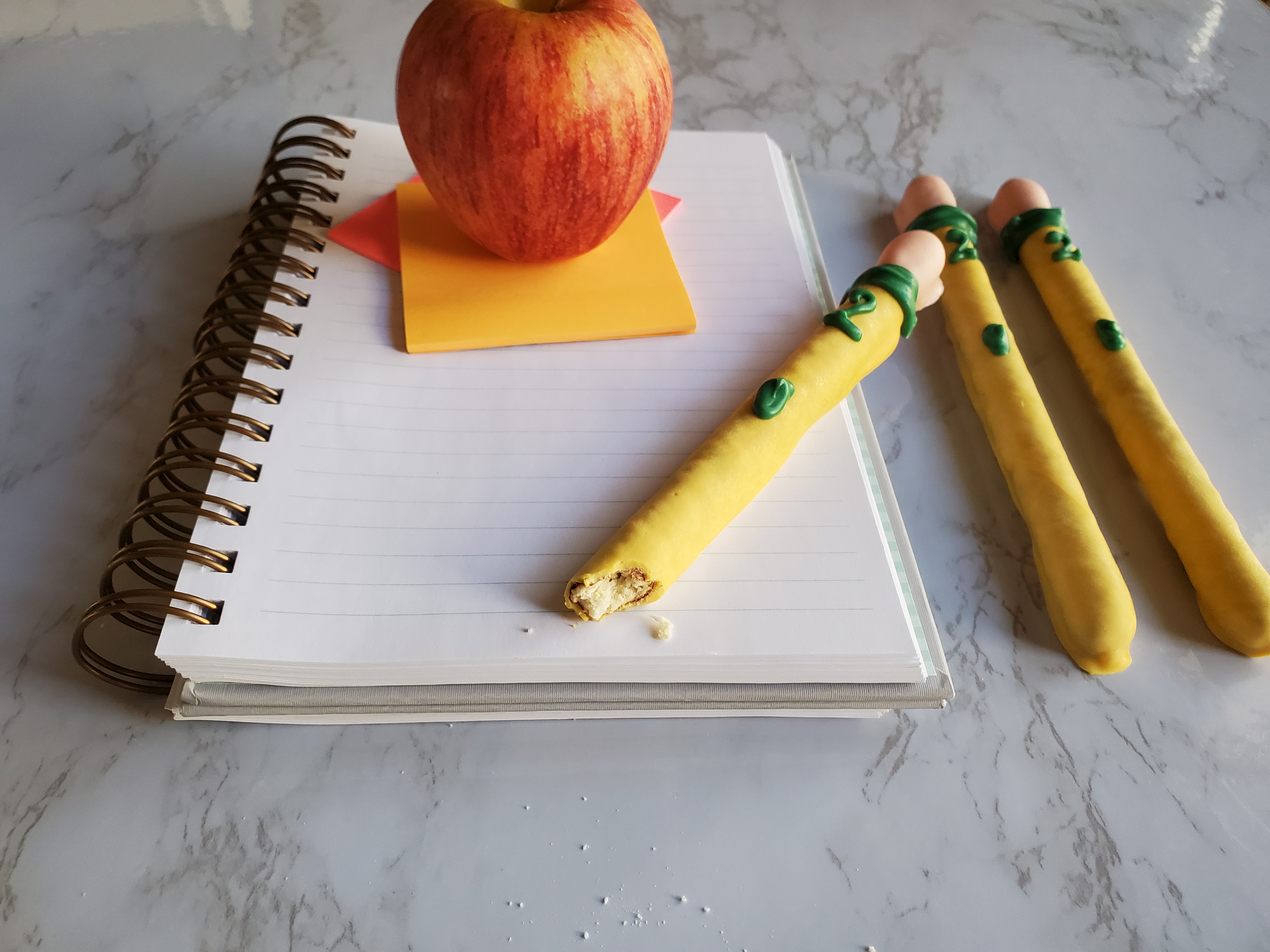 A notebook with bright pink and orange post-it notes, topped with a school lunch apple. A chocolate covered pencil pretzel sits on top of the notebook with a bite out of it, with two others to the right.