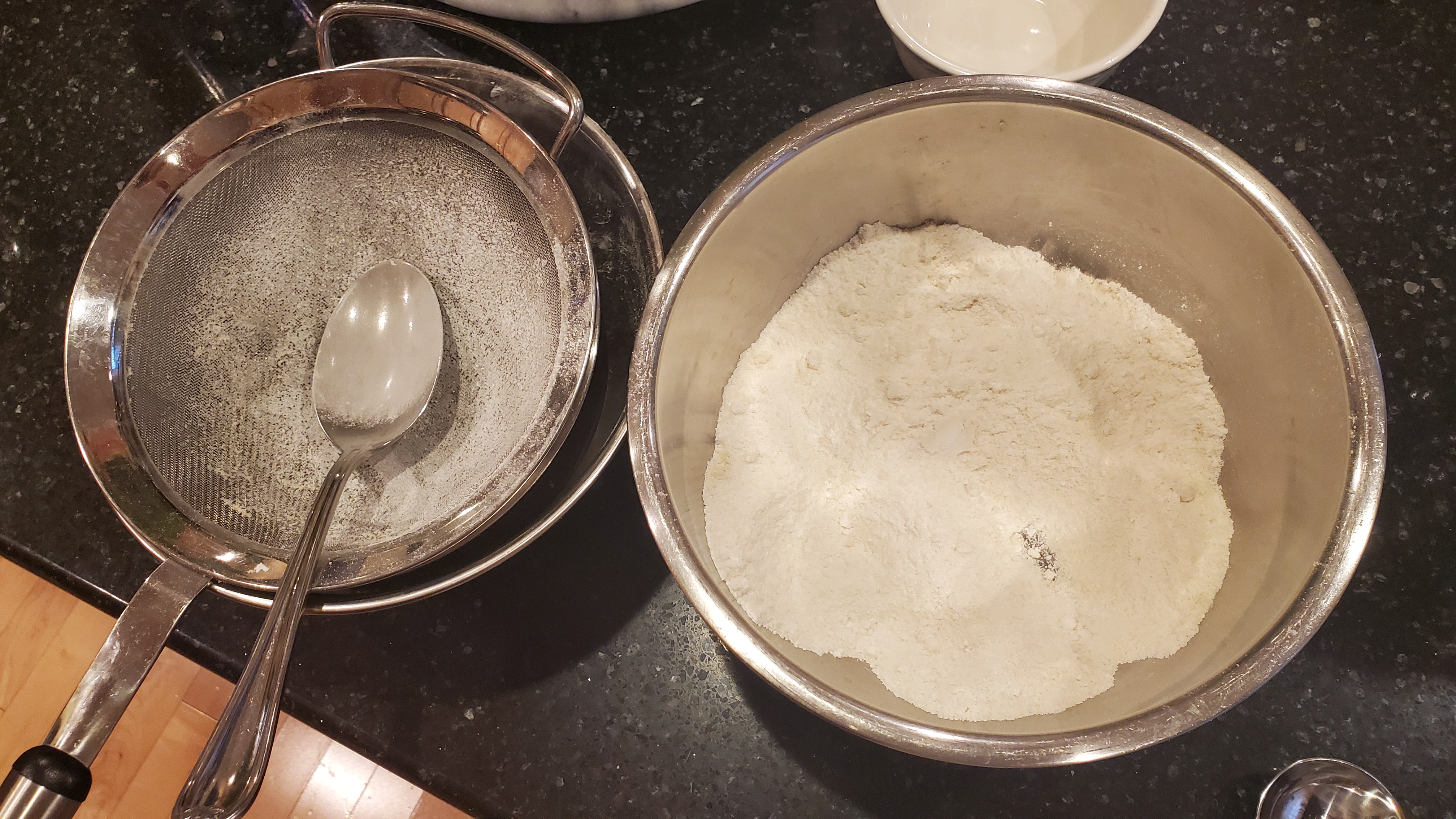 Powdered sugar and almond flour being sifted from a mesh strainer to a medium stainless steel bowl.