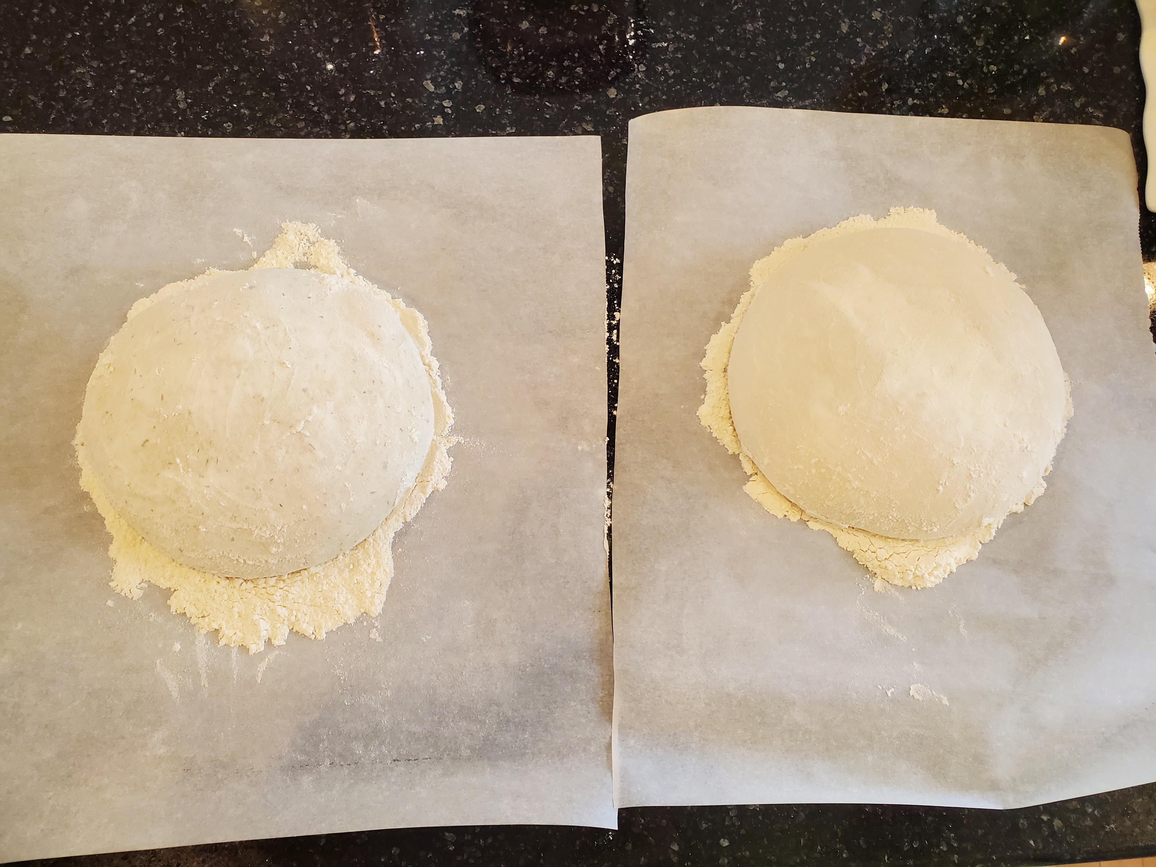 Two balls of sourdough rising on floured parchment paper on a black granite coutertop.