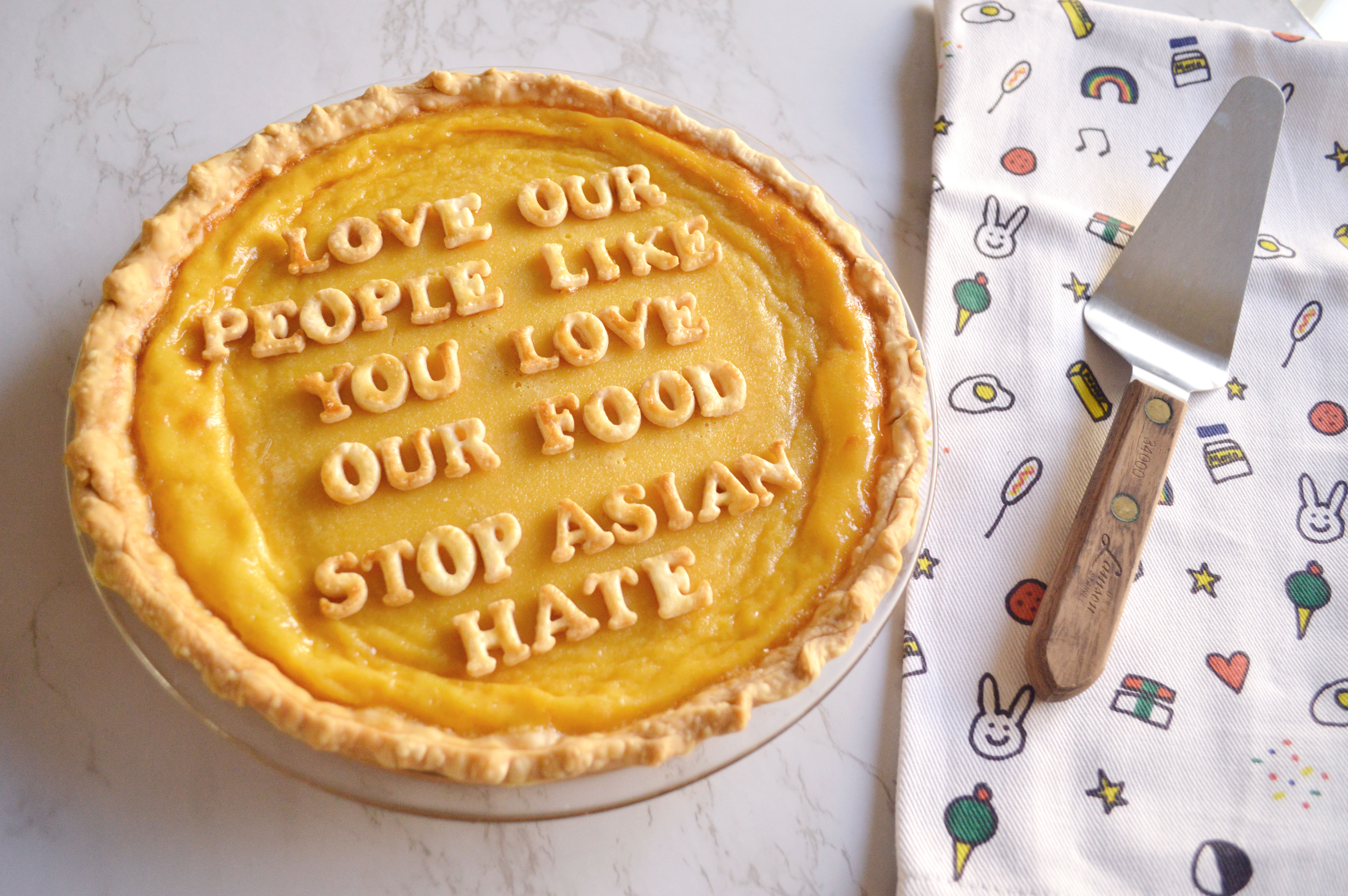 A giant egg tart sits in the lower left corner with pie crust letters spelling out "Love Our People Like You Love Our Food". It sits on a Molly Yeh No Kid Hungry dish towel with a pie turner on it's righthand side.