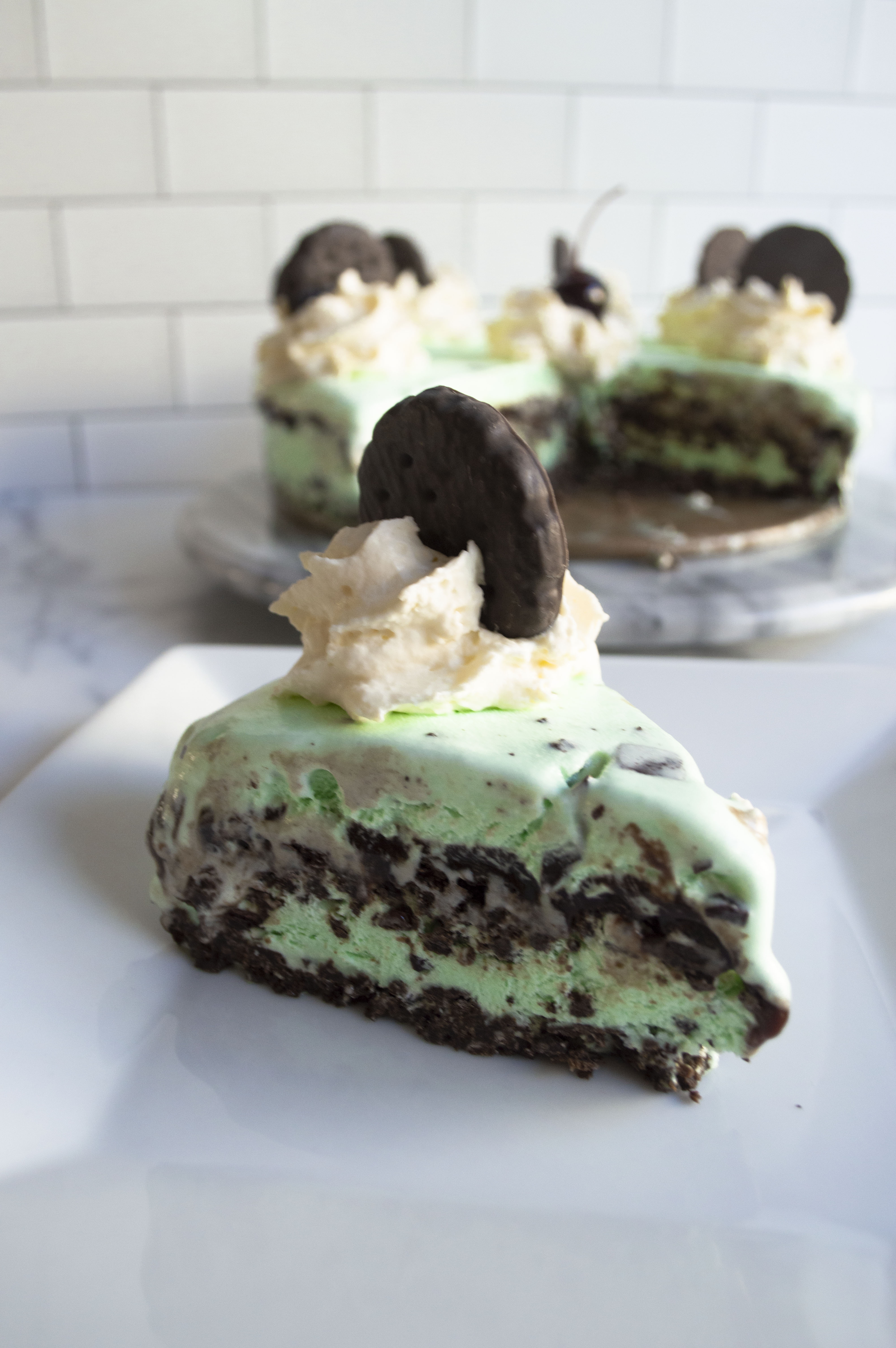 In the foreground sits a melty piece of ice cream cake. It is mint green with layers of cookie crust and fudge layered in, with whipped cream and another thin mint cookie on top. In the background against a subway tile backsplash, you can see the rest of the full cake.