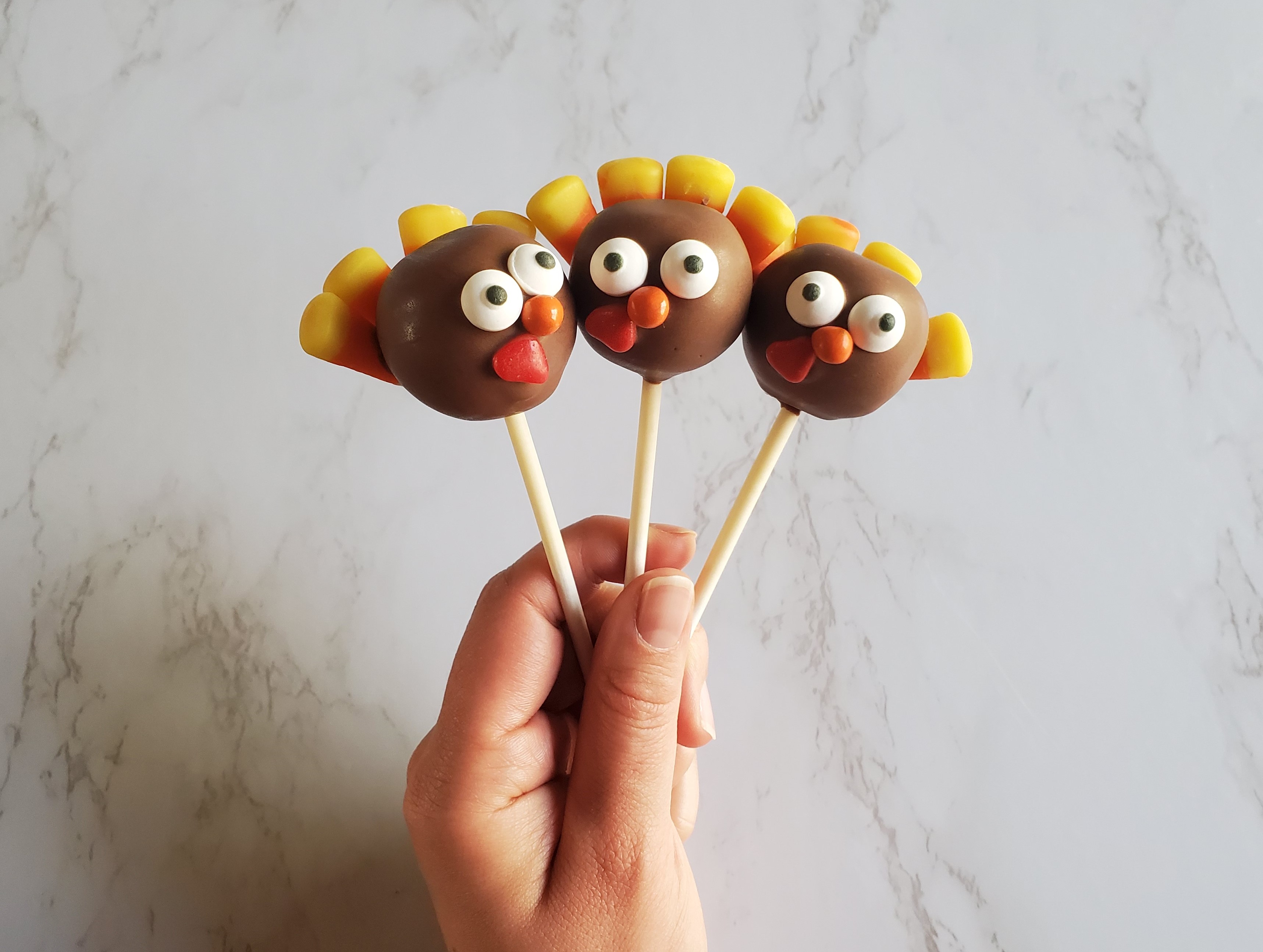 A hand holds up three cake pops decorated with candies including candy corn feathers to looks like turkeys in front of a marble background.