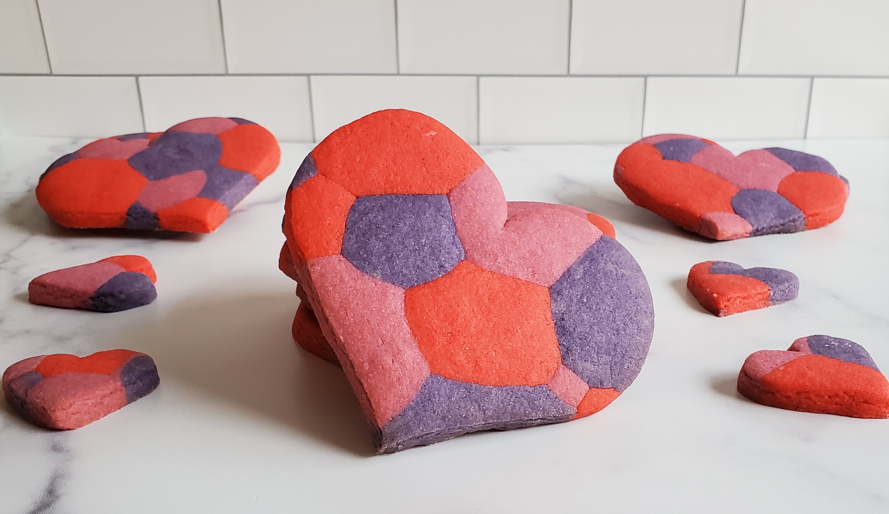 A large heart shaped sugar cookie with a marbled/mosaic pattern sits resting against a stack of more of the same cookies. On the sides we see slightly smaller heart shaped cookies of the same pattern. There are more large cookies in the back slightly propped up. All of this is on a white marble countertop against a subway tile backsplash.