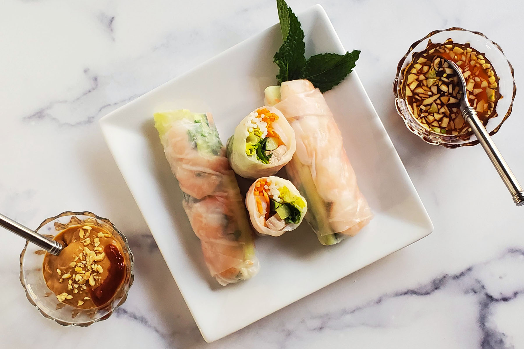 There are three Vietnamese Spring Rolls on a square, white plate. The roll in the middle is cut in half so we can see the shrimp, vegetables, pork and noodle inside. The plate is garnished with a sprig of mint leaf. On the diagonal sides, we see a peanut dipping sauce, sprinkled with crushed peanuts and sriracha on the lower left and a lighter brown colored sauce with floating pieces of mince garlic on the upper right. This is all on top of a white marble countertop.