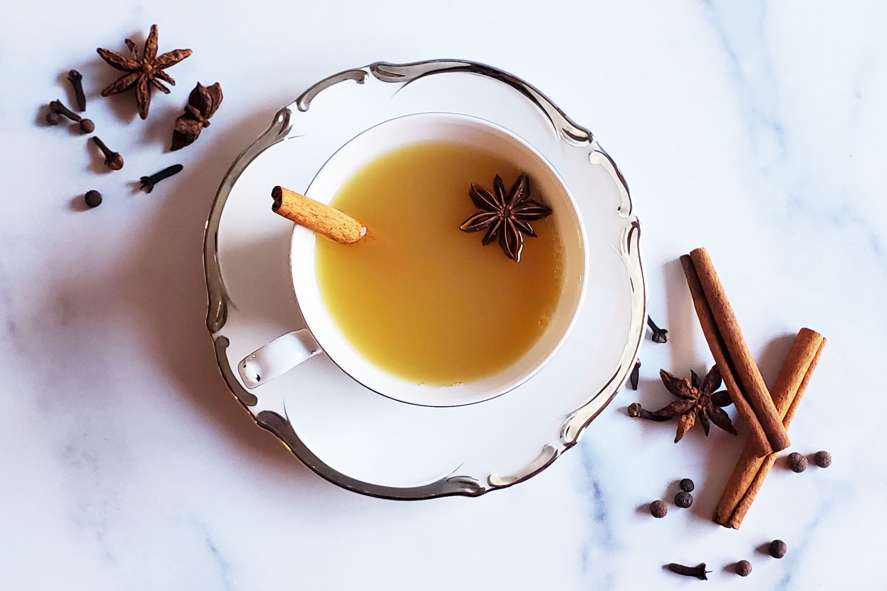 An antique teacup on a saucer, filled with wassail, a cinnamon stick and one star anise. It is in a marble counter, surrounding the cup are more spices, cinnamon sticks, clove, allspice berries and star anise.