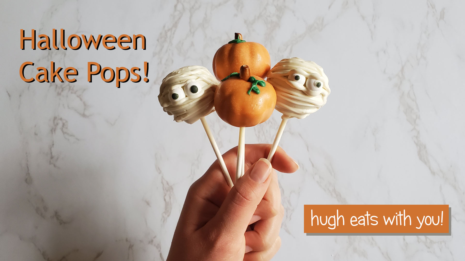 A hand holds up mummy and pumpkin cake pops in front of a white marble background. The photo reads, "Halloween Cake Pops!" in the upper left corner and "hugh eats with you" in the bottom right corner.