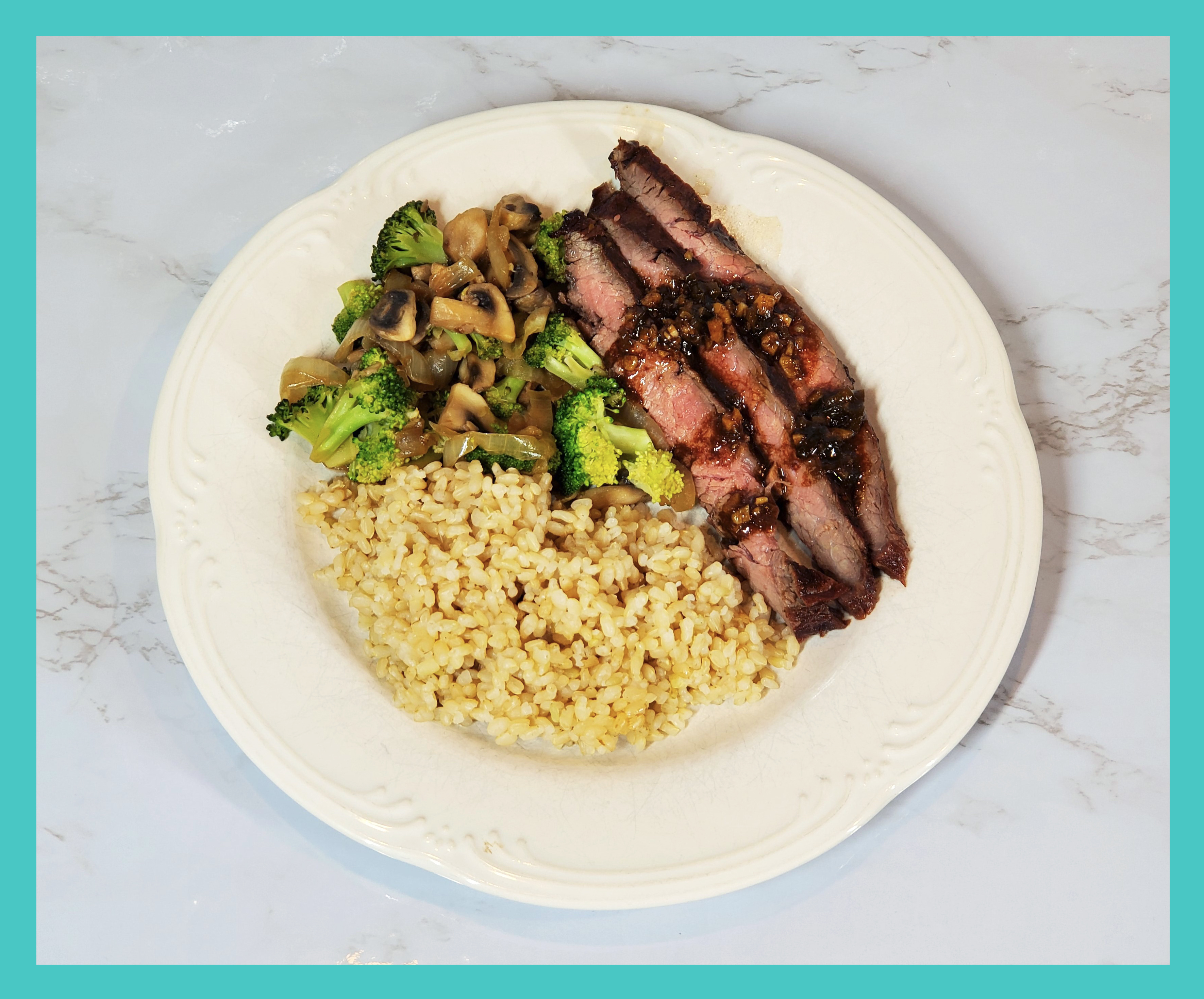 Flank Steak drizzled in sauce on a white plate with short grain brown rice, broccoli/mushrooms/onions. On a white marble background.