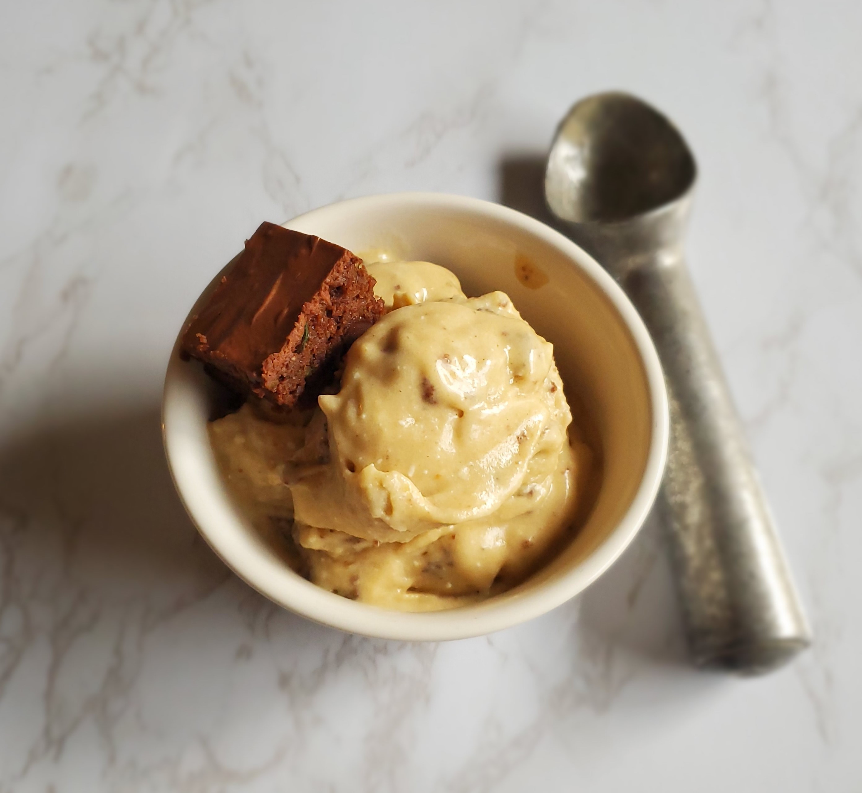 Vegan Peanut Butter Ice Cream in on white bowl on white marble with a chunk of brownie and a silver spoon.