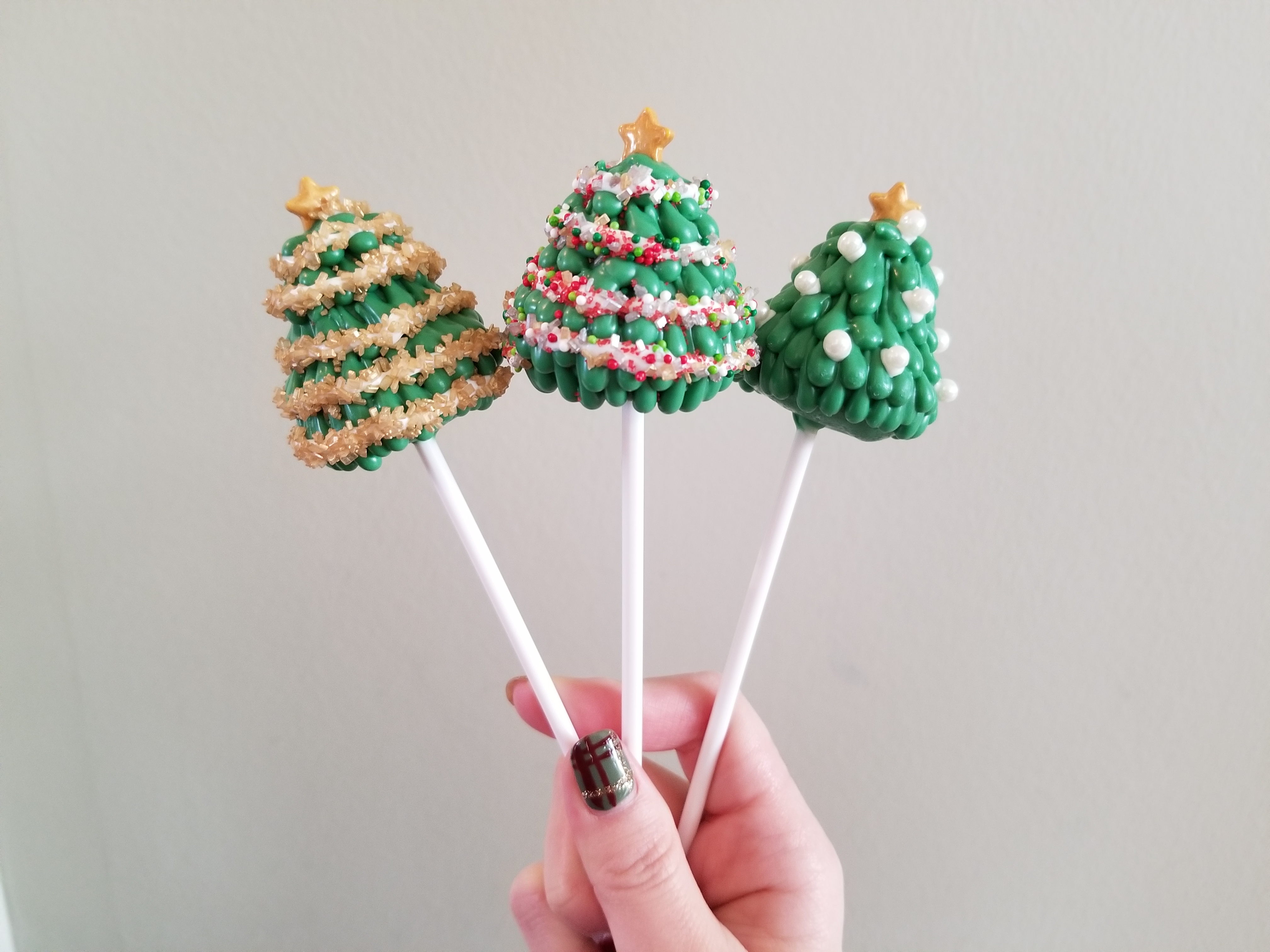 In front of a white background a woman with green painted nails holds three Christmas tree cake pops. They are decorated with sprinkles to resemble a string of light around them. Starting from the left on has gold sprinkles, red/green/white nonpareils and finally individually placed pearl sprinkles.