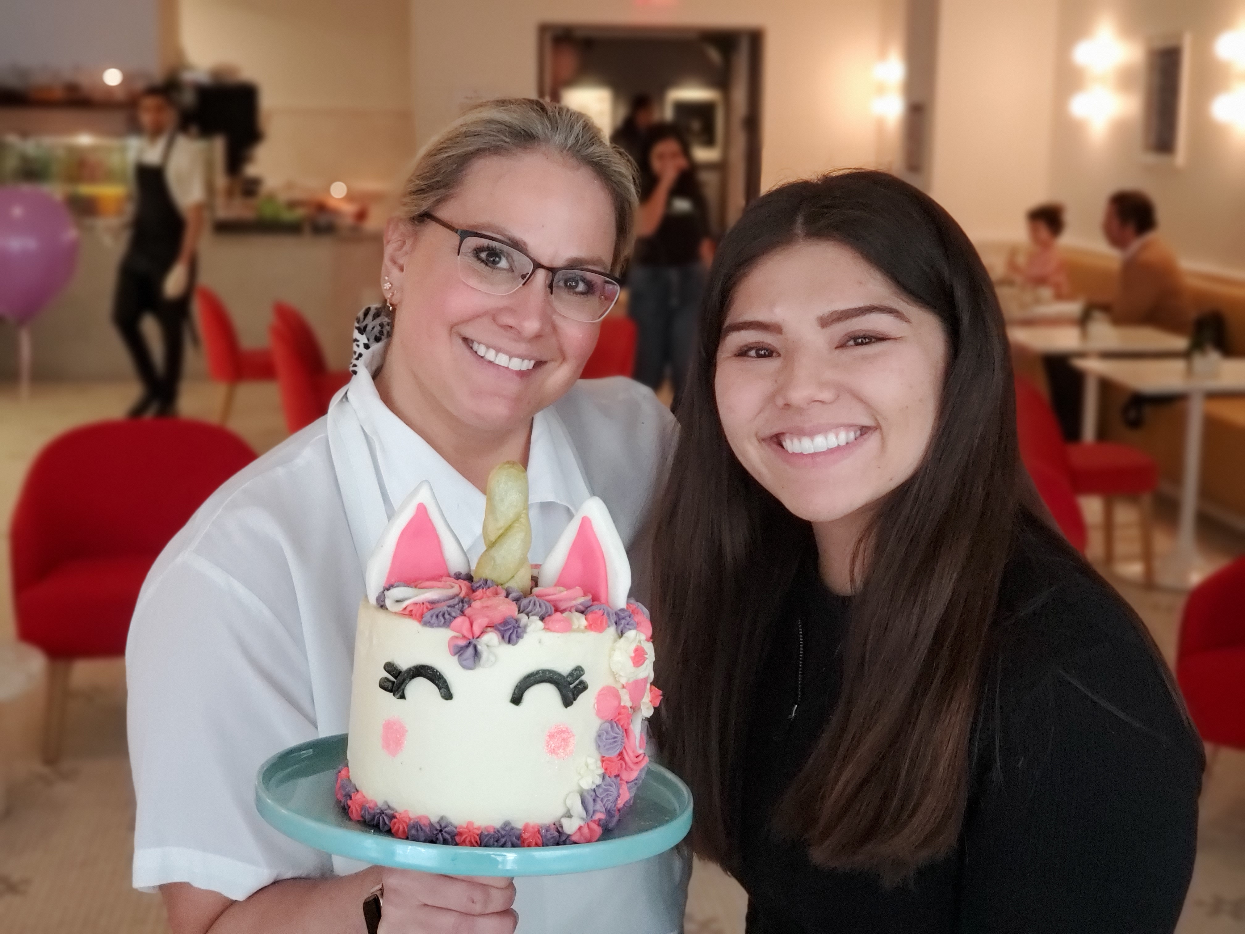 Two women holding a unicorn cake with pink and purple flowered hair and a gold horn. The woman on the left is blonde, her hair pulled back with a white and black polka-dotted bow. Wearing black glasses and a short-sleeved white chef's coat. The woman on the right (me) is a brunette, in an all black peplum zip up sweater. Her hair is down, she is smiling. In the background you can see blurred red chairs and families sitting at cafe table.