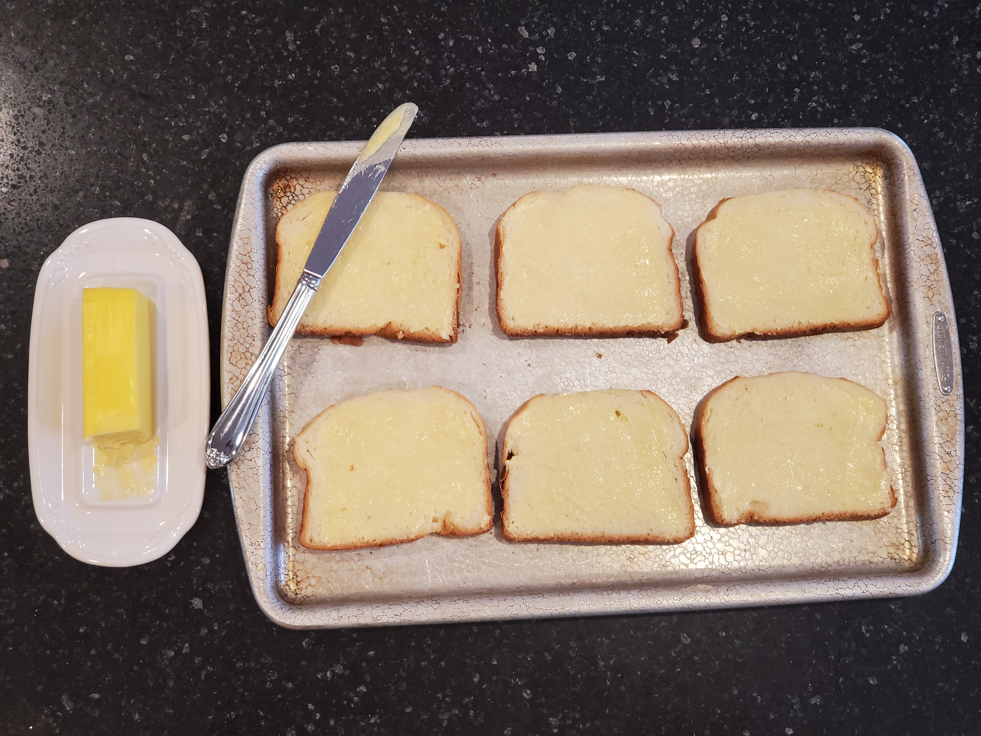 Six slices of brioche sit on a baking sheet buttered, a dish with softened butter and a butter knife sit beside. All on a black granite countertop.
