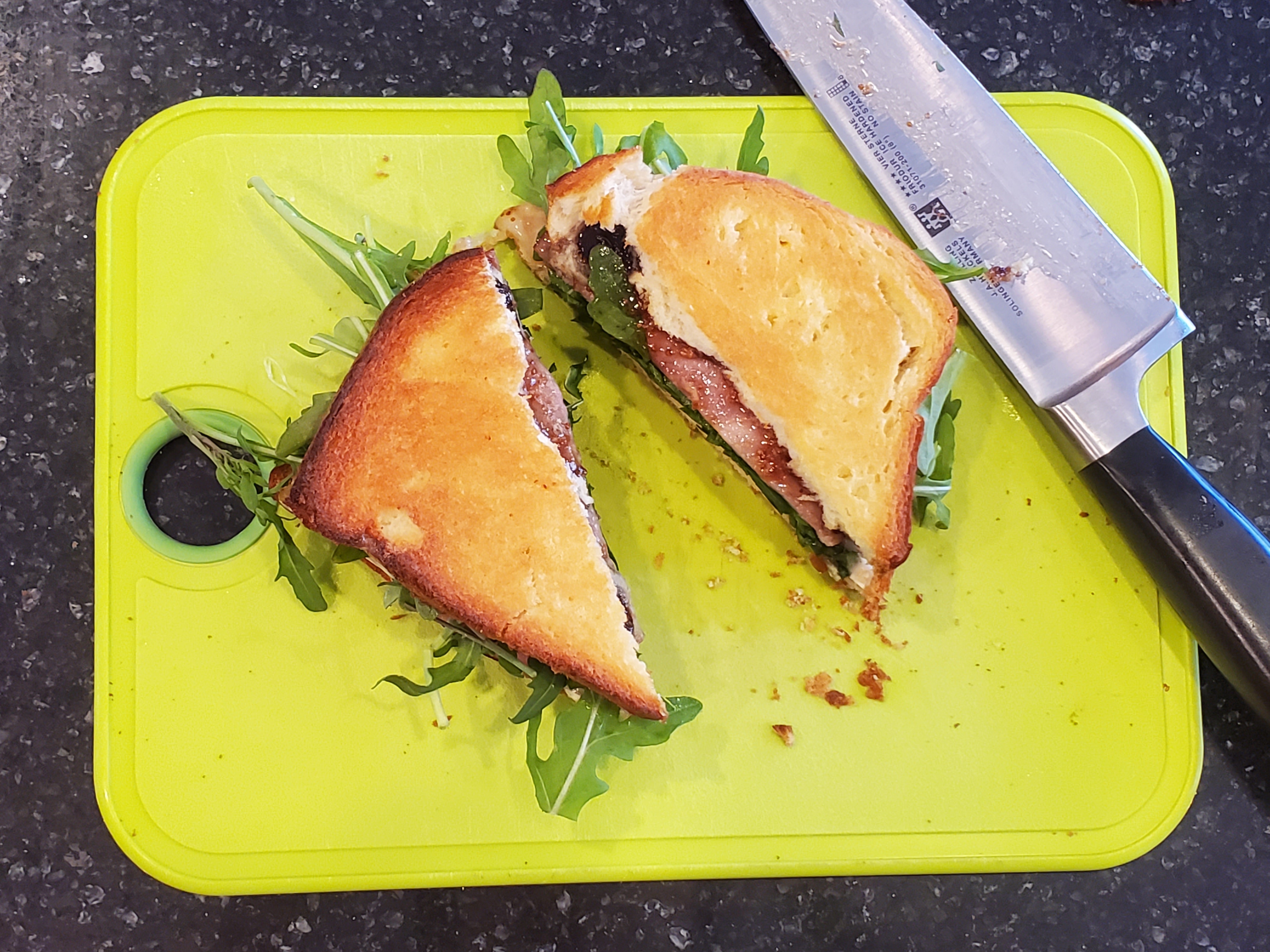 The brie/fig/arugula grilled cheese sits a top the bright lime green cutting board, cut diagonally in half. A sharp knife sits at the right.