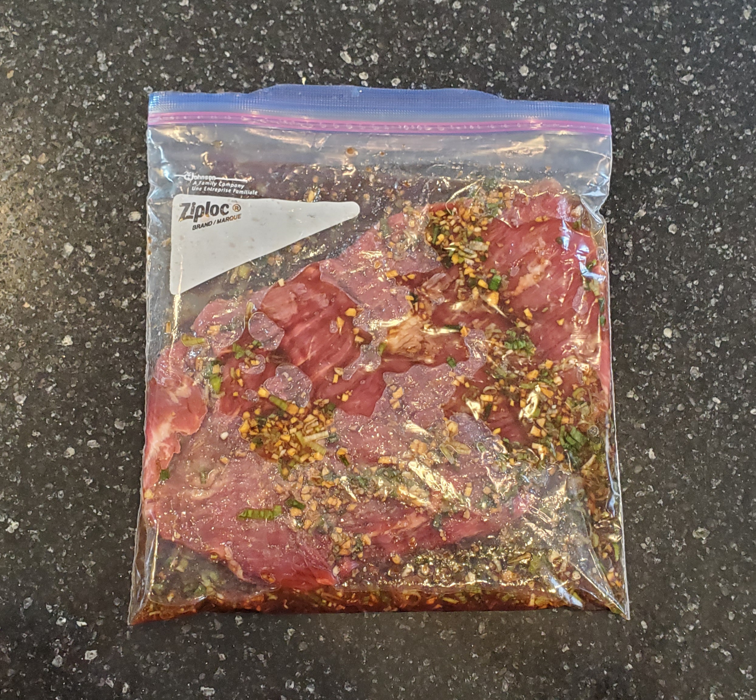 Marinated flank steak in a gallon, plastic, Ziploc bag on a black granite countertop