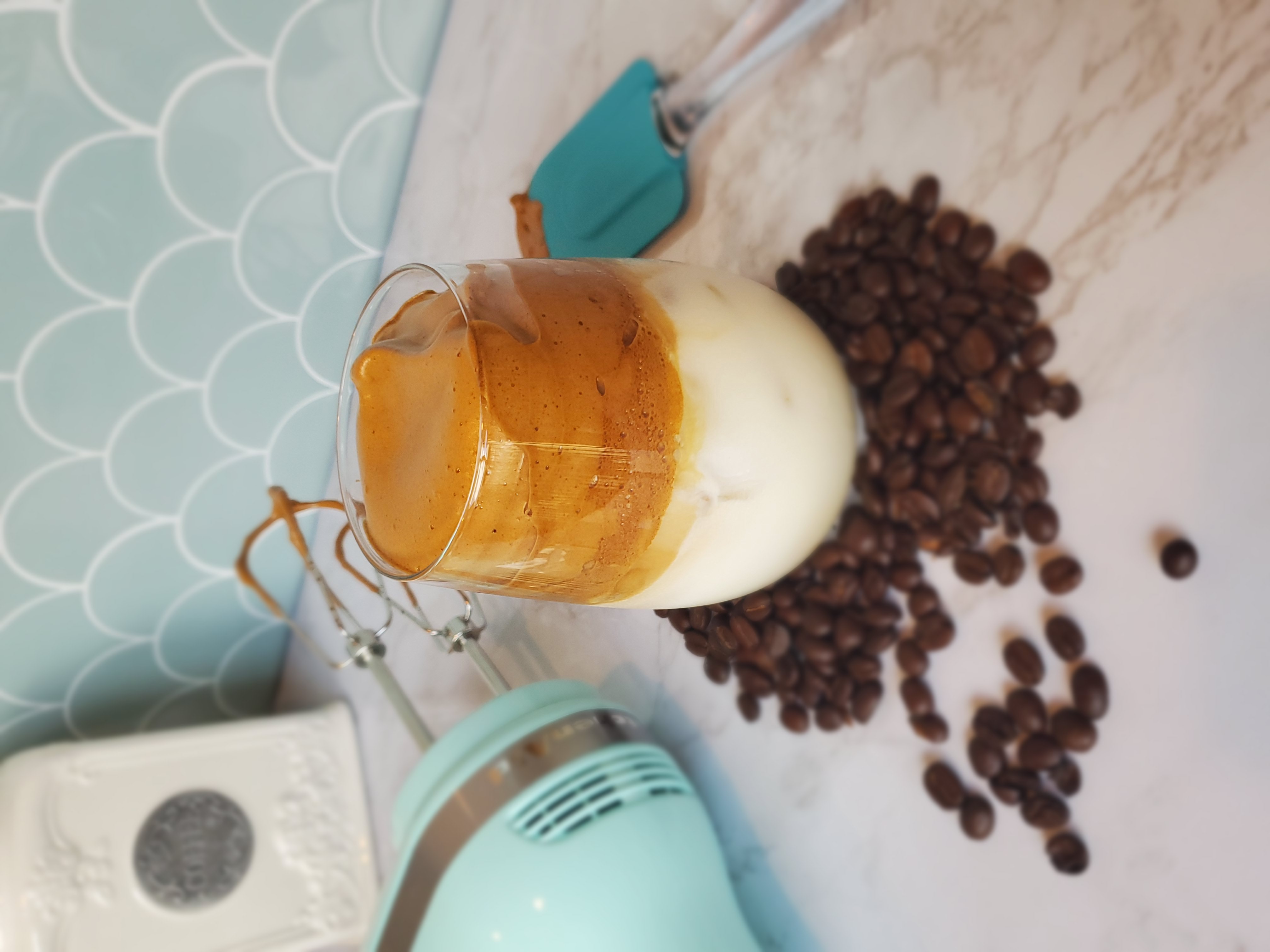 A Dalgona coffee sitting in coffee beans and a next to a dirty, turquoise kitchen aid hand mixer and spatula. 