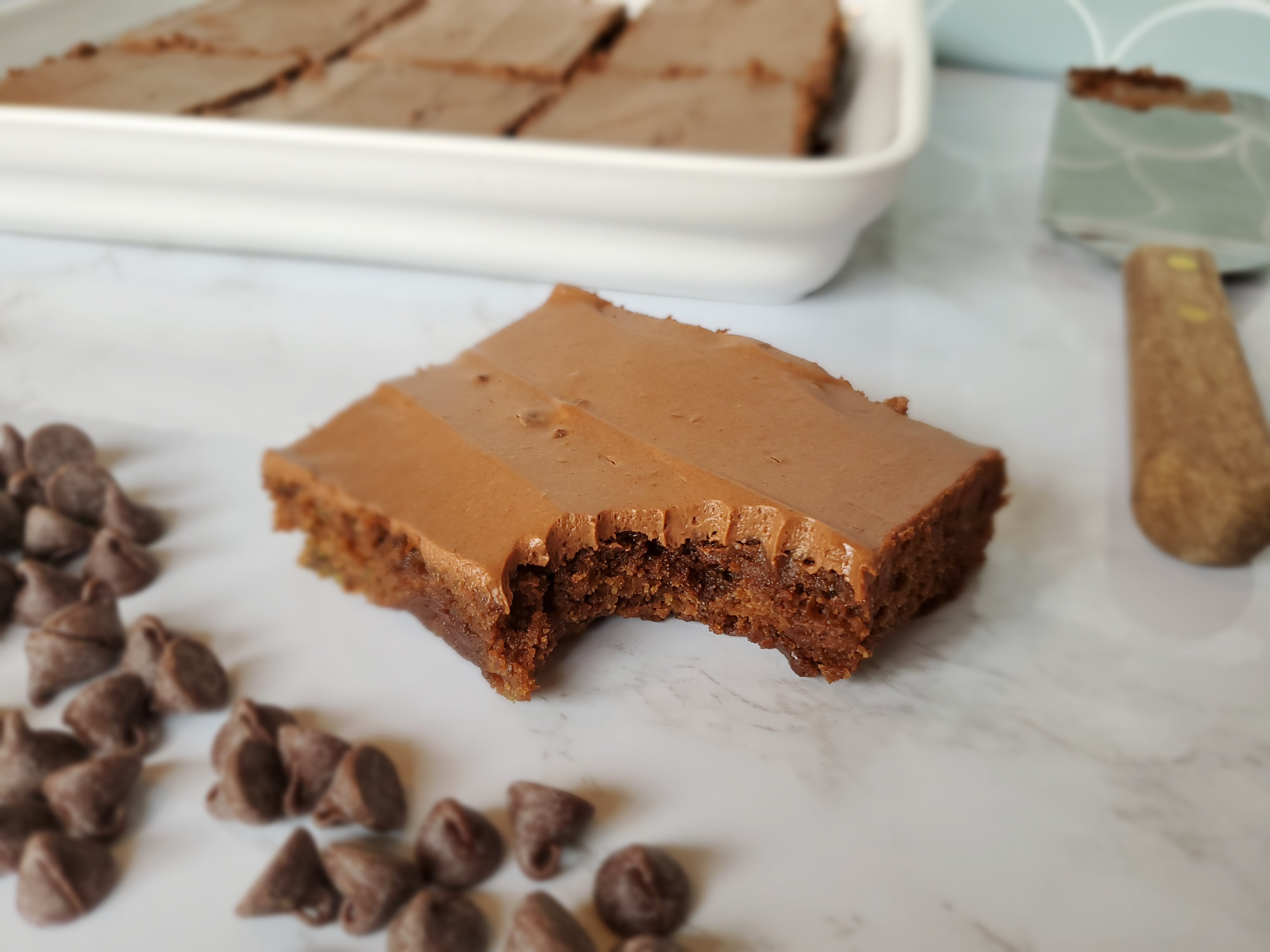 Vegan zucchini brownie surrounded by chocolate chips, a platter of more brownies sits behind it as well as a spatula to it's right.