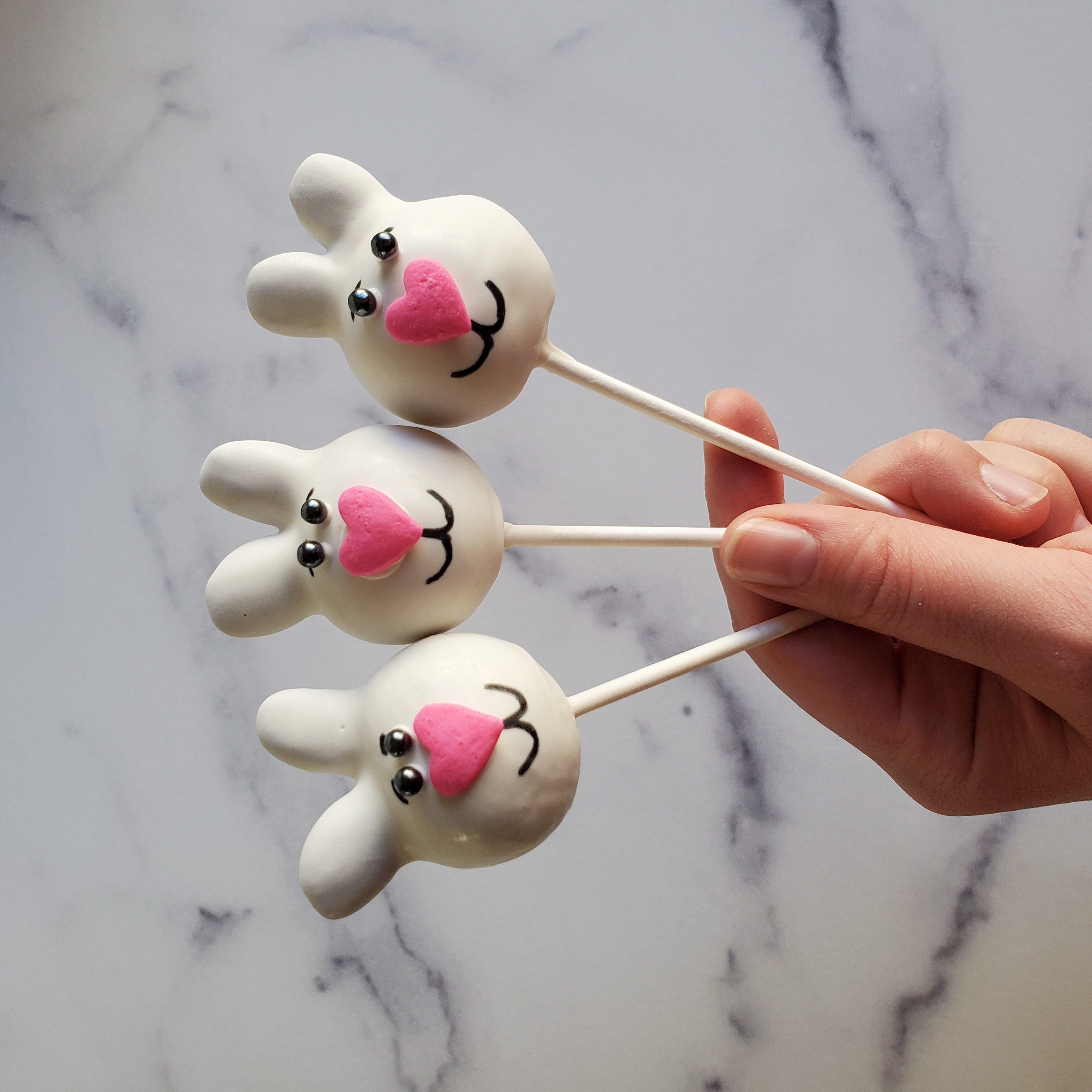 A person (me) holds three white Easter Bunny cake pops in her left hand. The bunnies have black sprinkles for eyes, a large pink, sprinkle heart nose and a mouth drawn on with a black food safe marker.