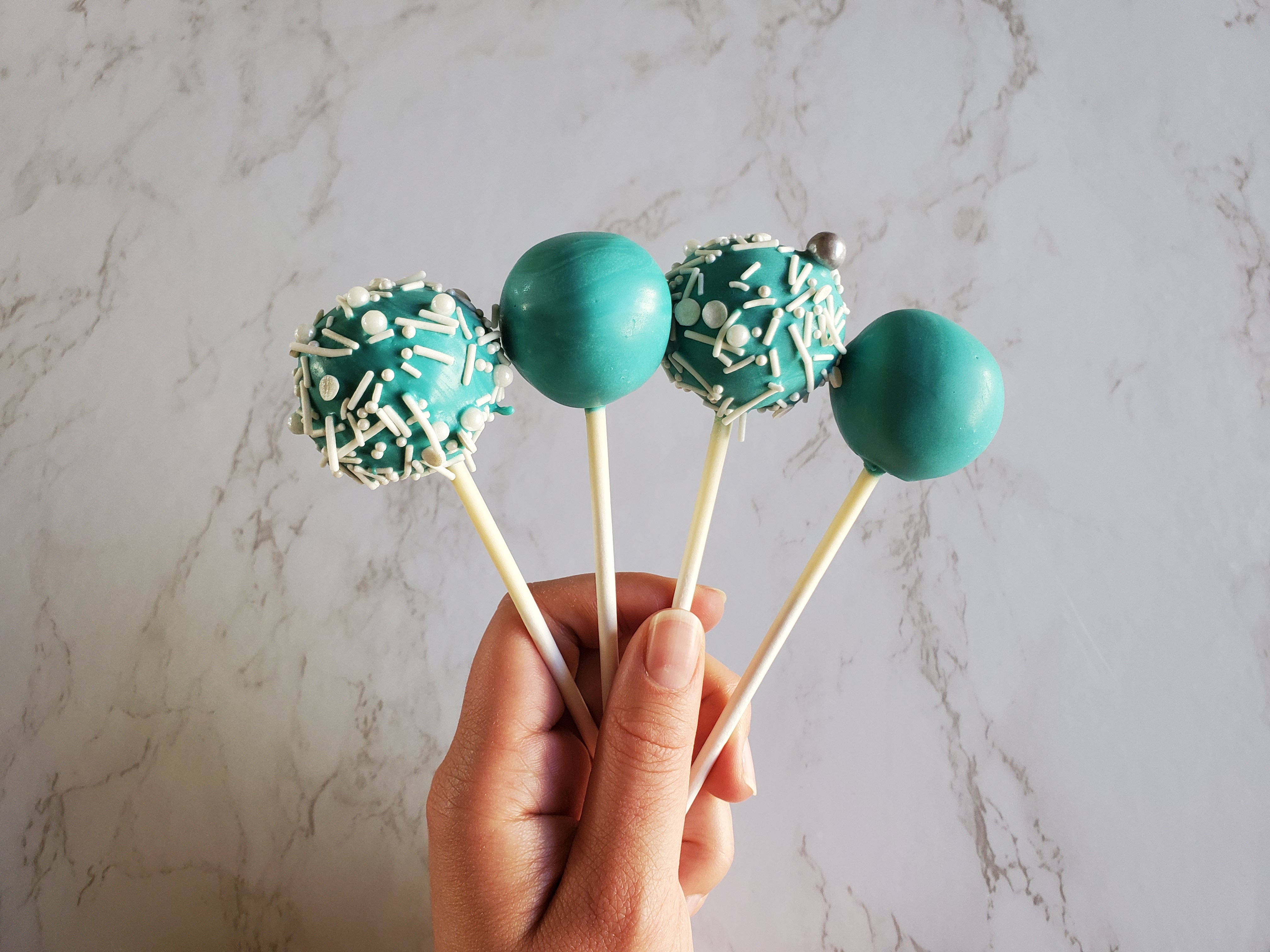 Turquoise cake pops with white and shiny round, gray sprinkles in a hand in front of a marble background.