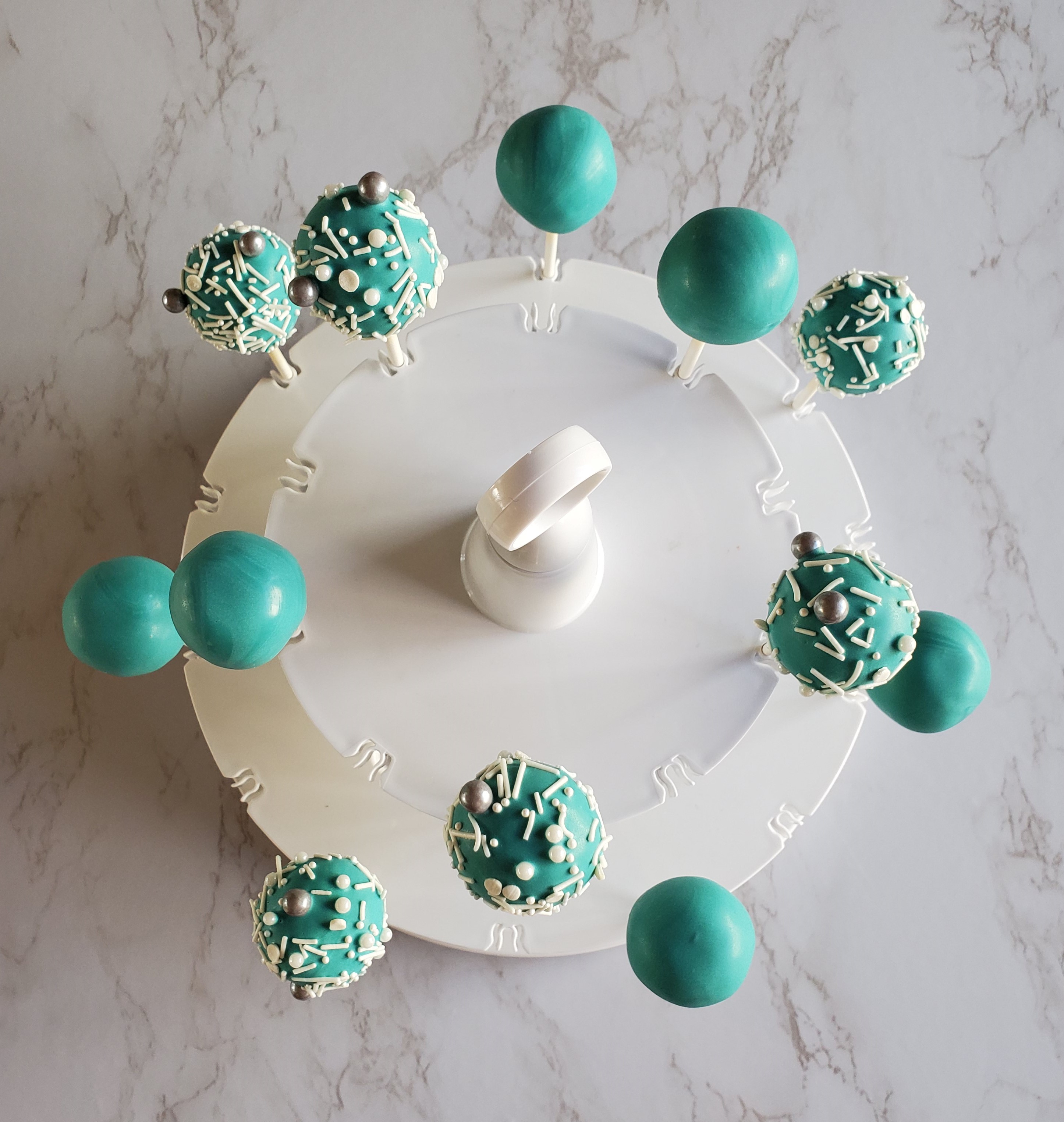 Turquoise cake pops with white and shiny round, gray sprinkles in front of a marble background in a cake pop stand.