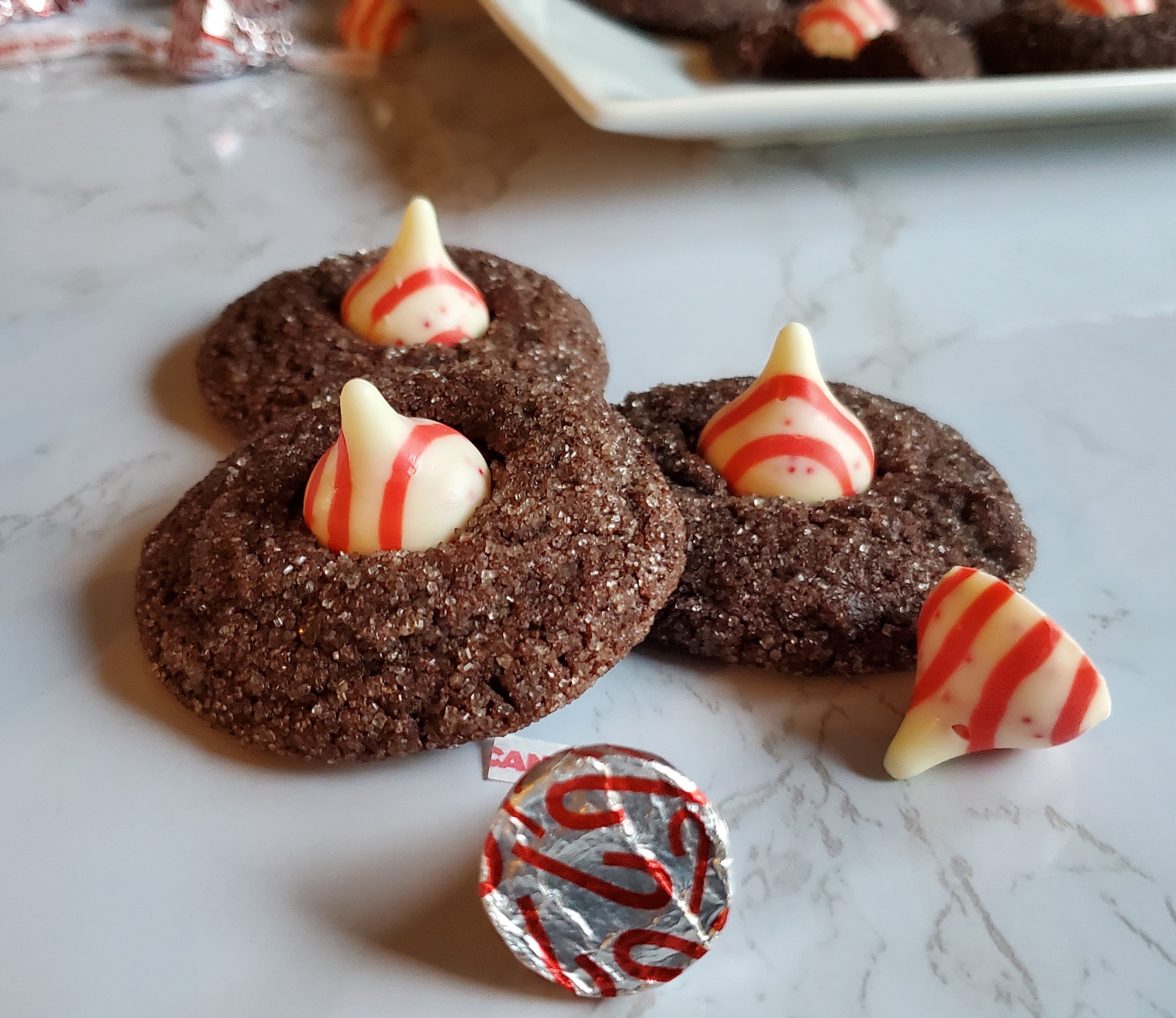 Chocolate cookies, rolled in sugar with a white chocolate candy cane hershey's kiss in the top. There are three stacked around each other with candies and wrappers around them.