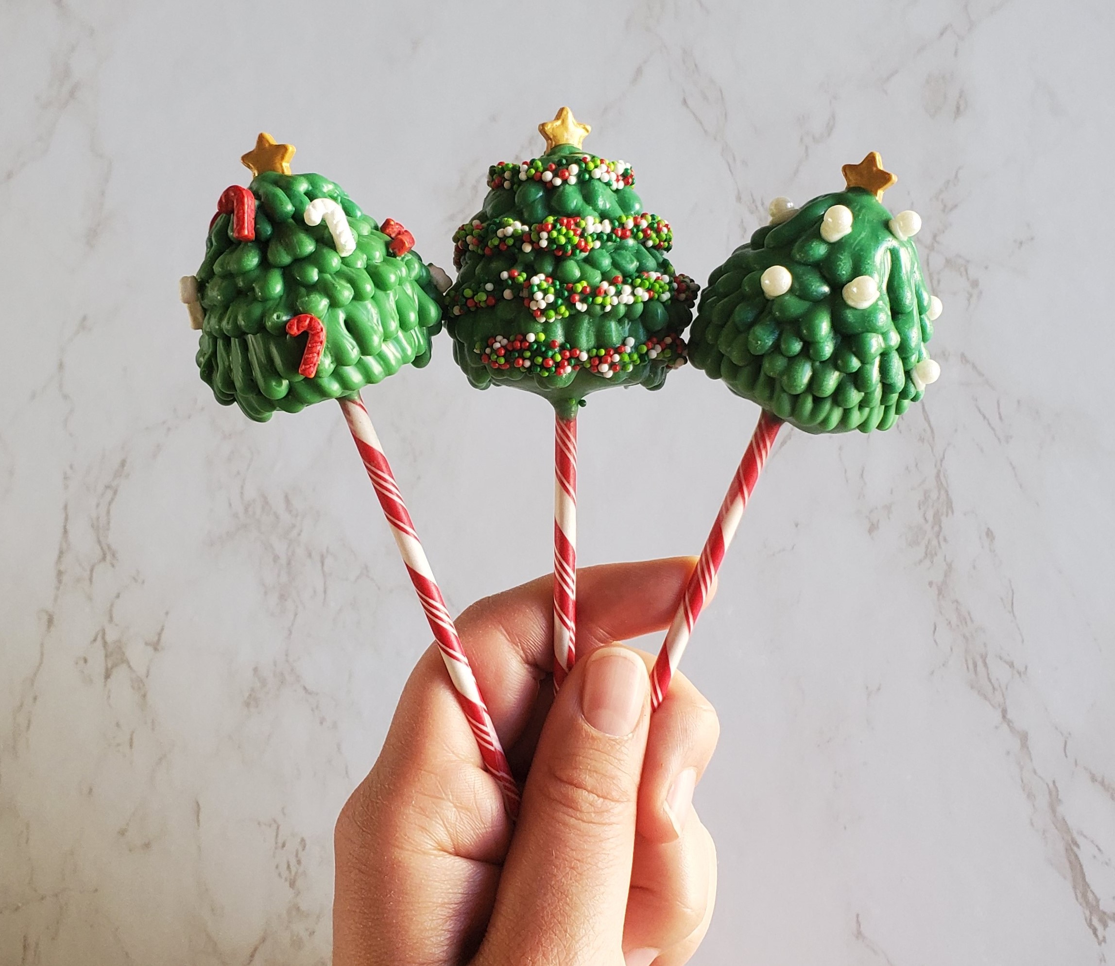 Three Christmas cake pops held in my left hand. The sticks are candy cane stripped and the trees have a star sprinkle on top, along with sprinkles around the pop, to look like garland.