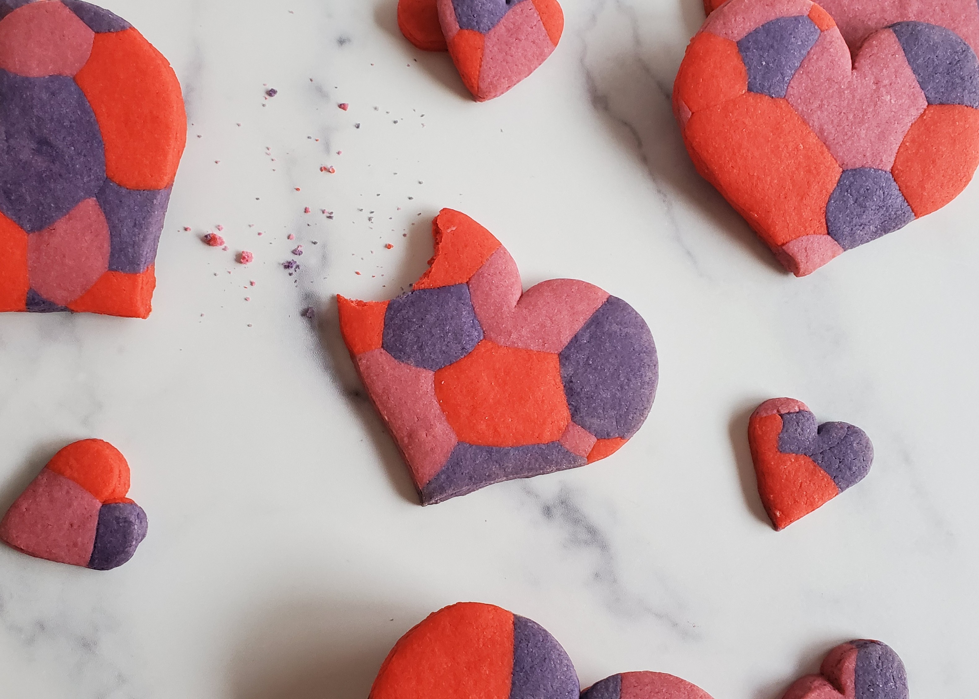 A multi-colored heart shaped cookie in pink red and purple with a big crumby bite out of it, surrounded by similar cookies on a white marble background.