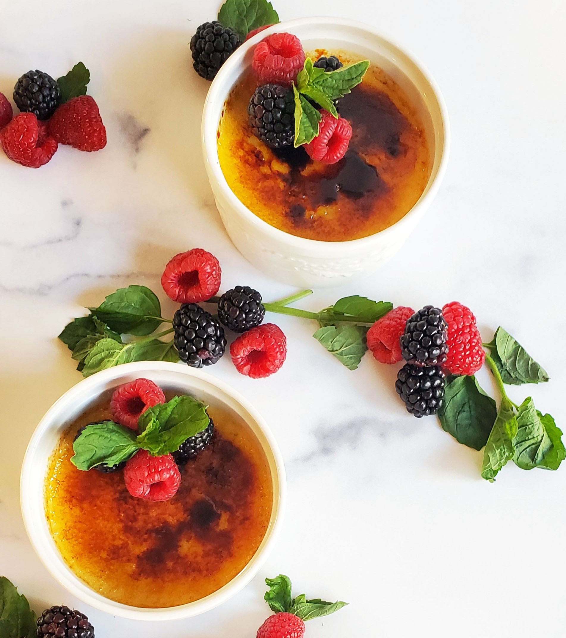 Two ramekins of creme brulee, surrounded by raspberries, blackberries and mint sprigs on a white marble background.