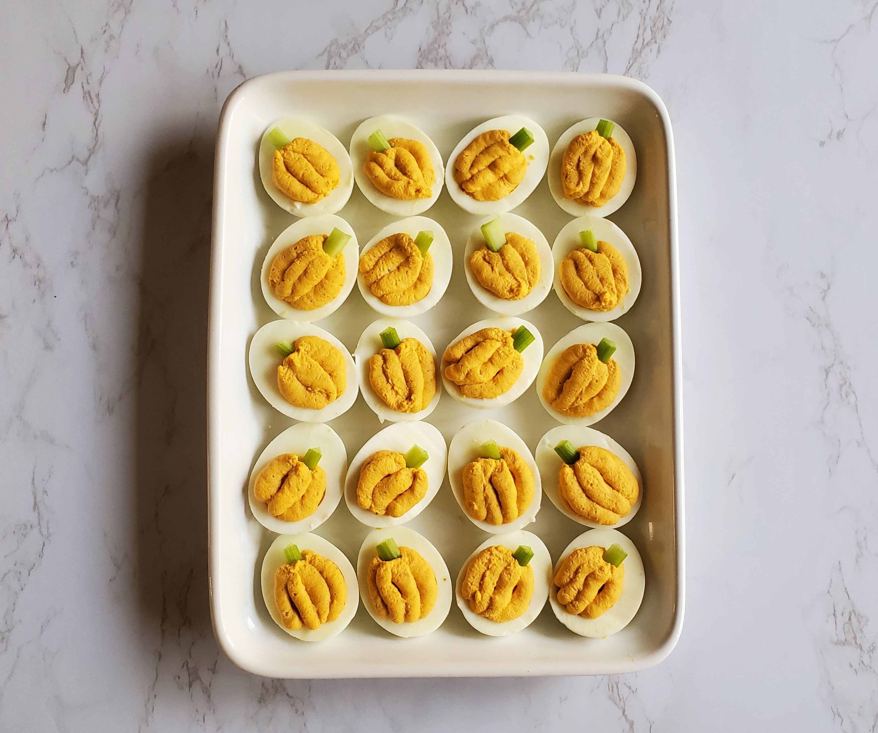 A white rectangular platter of devilled eggs, made to look like pumpkins (with celery stems), sits on top of a white marble countertop.