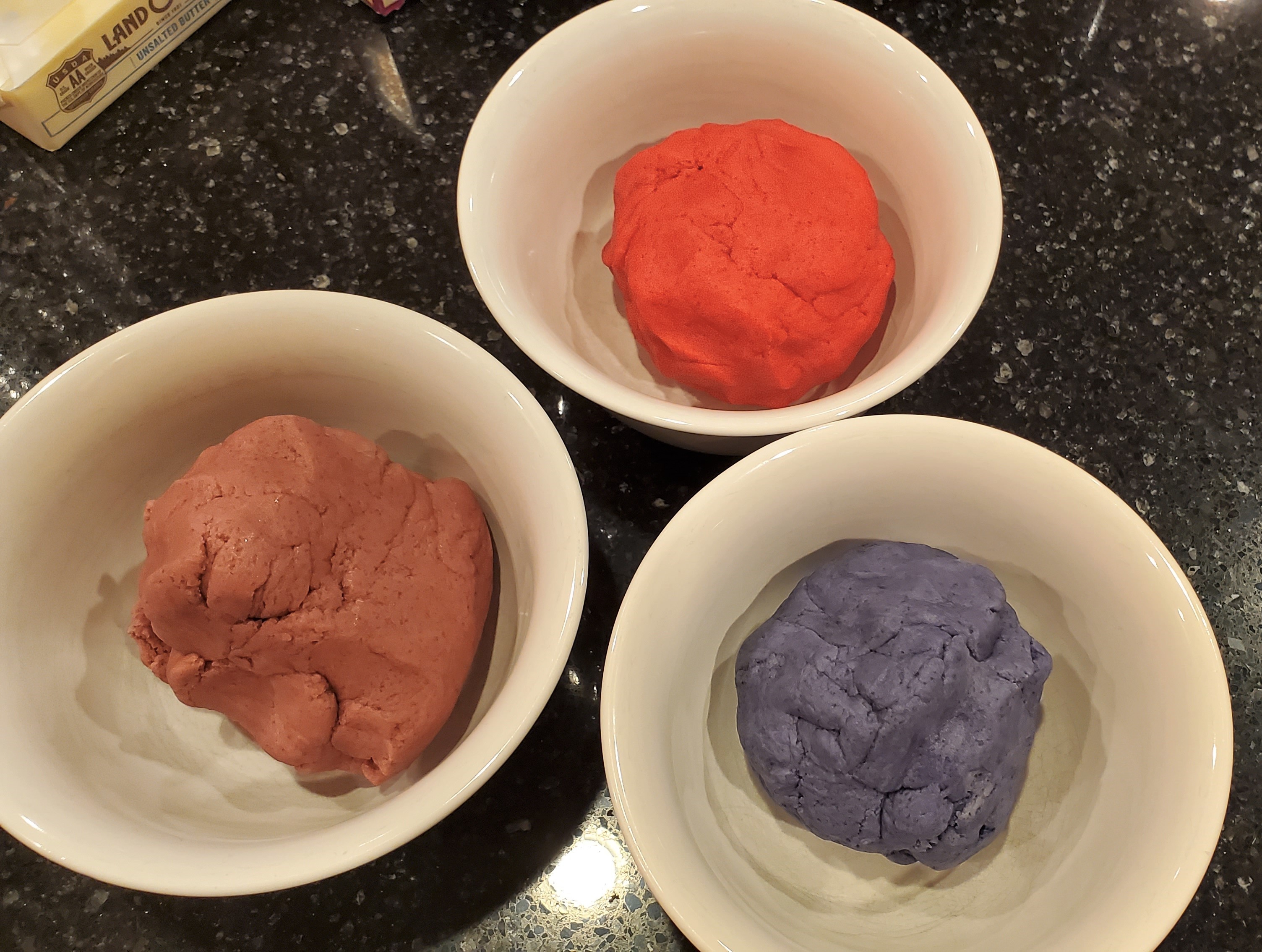 Three dough balls in pink, red and purple. Each are in individual white bowls on a black granite background. You can also see a partial stick of butter and some flour.