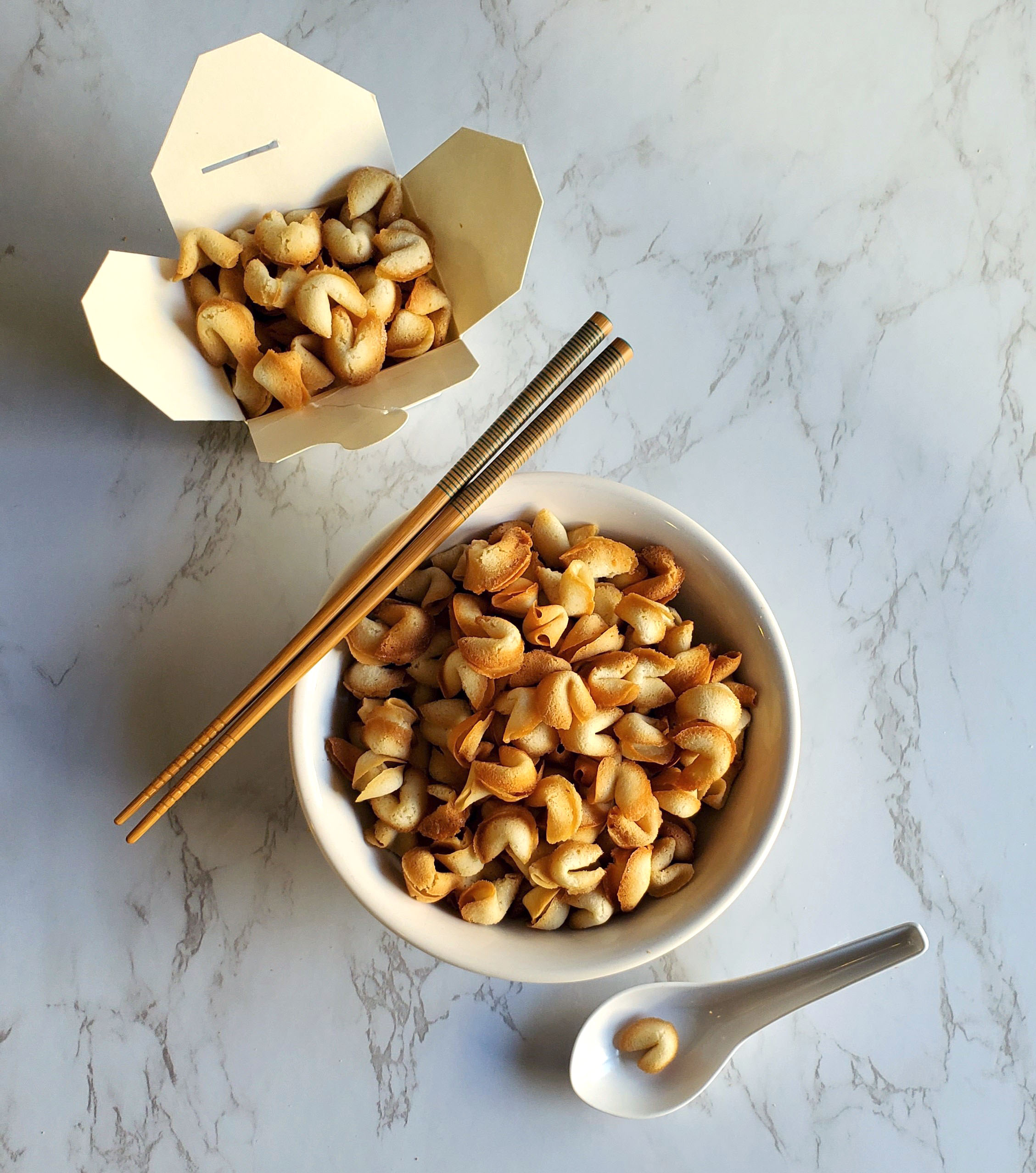 There is a white bowl full of "fortune cookie cereal". Resting on the bowl is a pair of chopsticks with a green lined pattern that wraps around the top third. In the upper left there is a small Chinese takeout box filled to the brim with fortune cookie cereal. All on a white marble countertop.