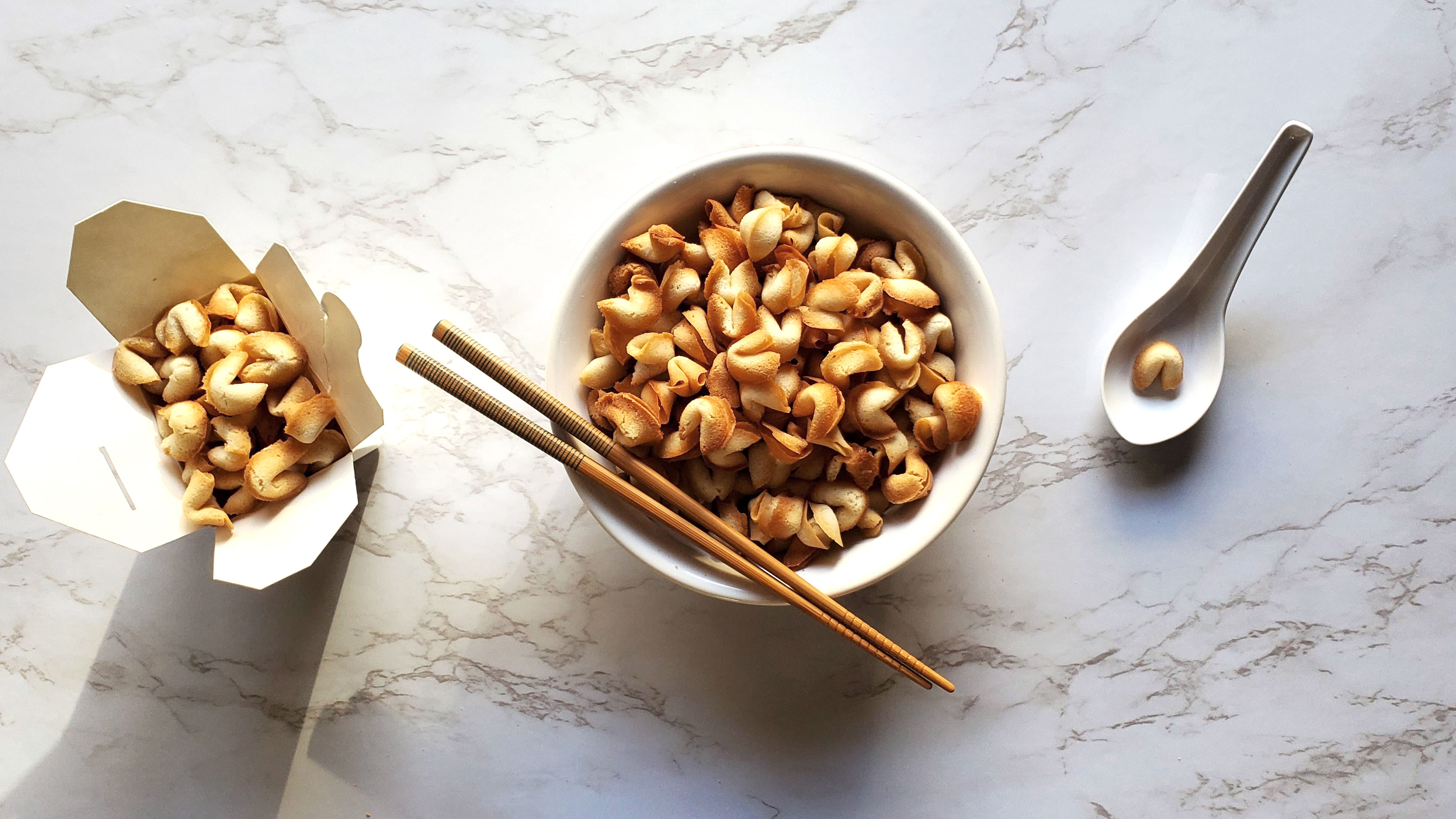 There is a white bowl full of "fortune cookie cereal". Resting on the bowl is a pair of chopsticks with a green lined pattern that wraps around the top third. In the upper left there is a small Chinese takeout box filled to the brim with fortune cookie cereal. All on a white marble countertop.