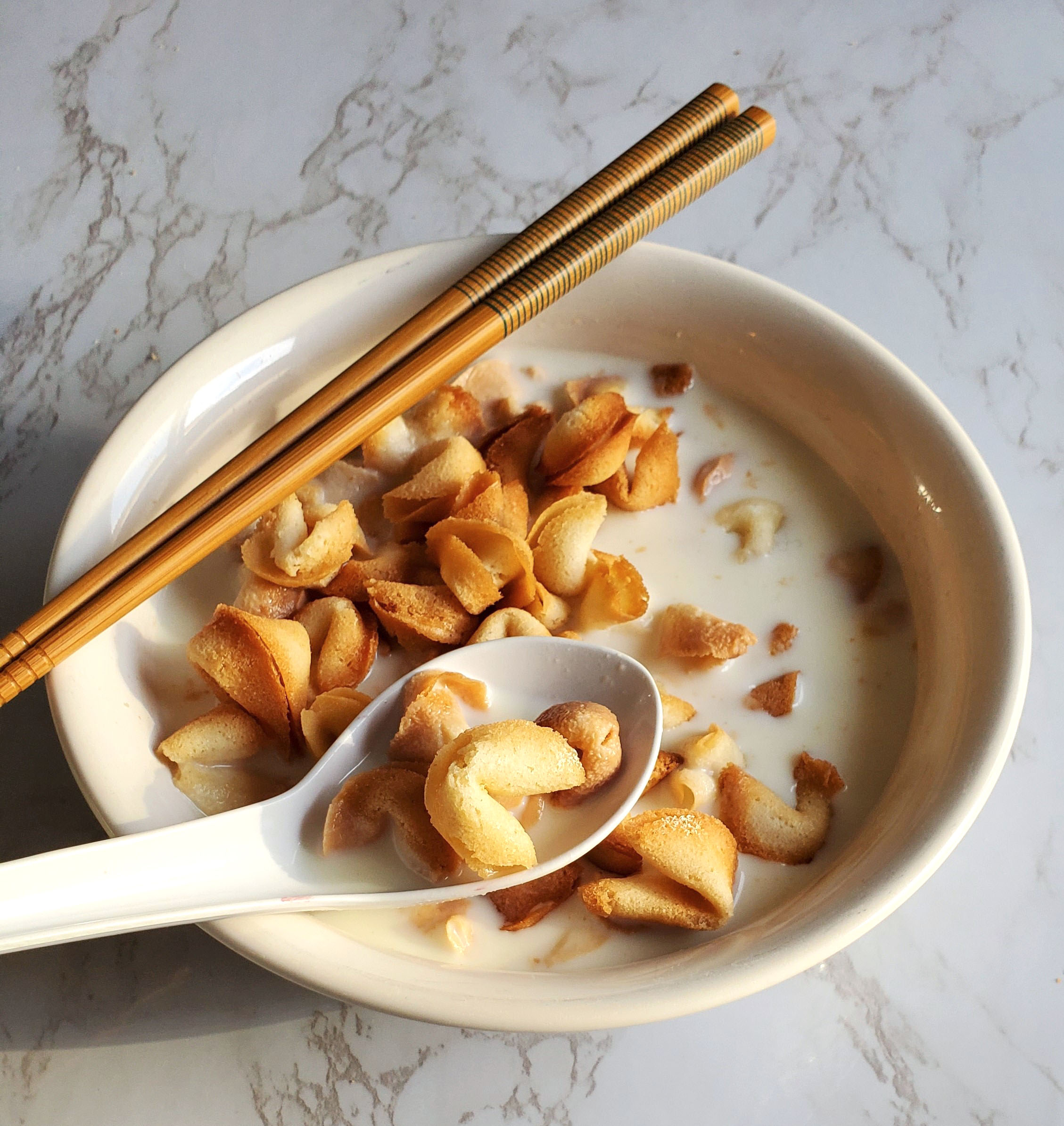 There is a white bowl, filled with milk and many tiny fortune cookies. Inside is a bowl filled with a scoop full of the cookies. Chopsticks with a green lined pattern rest diagonally on the upper left hand side of the bowl. The bowl sits on a white countertop.