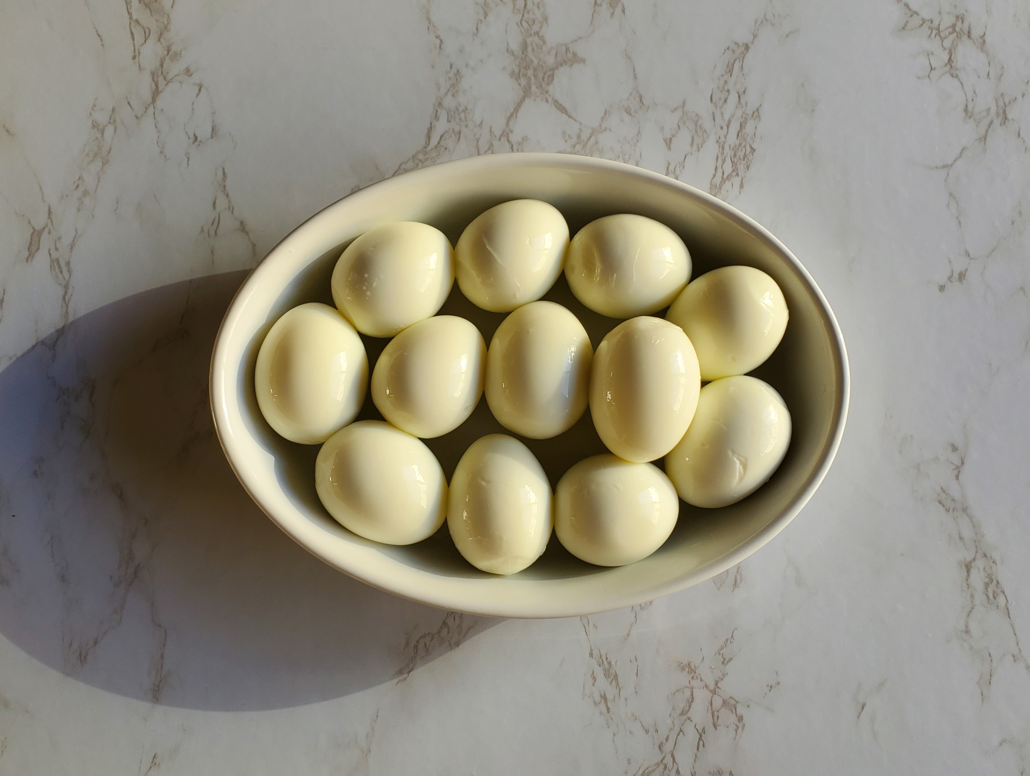 A white oval ceramic platter on a marble counter top filled with hard boiled eggs.