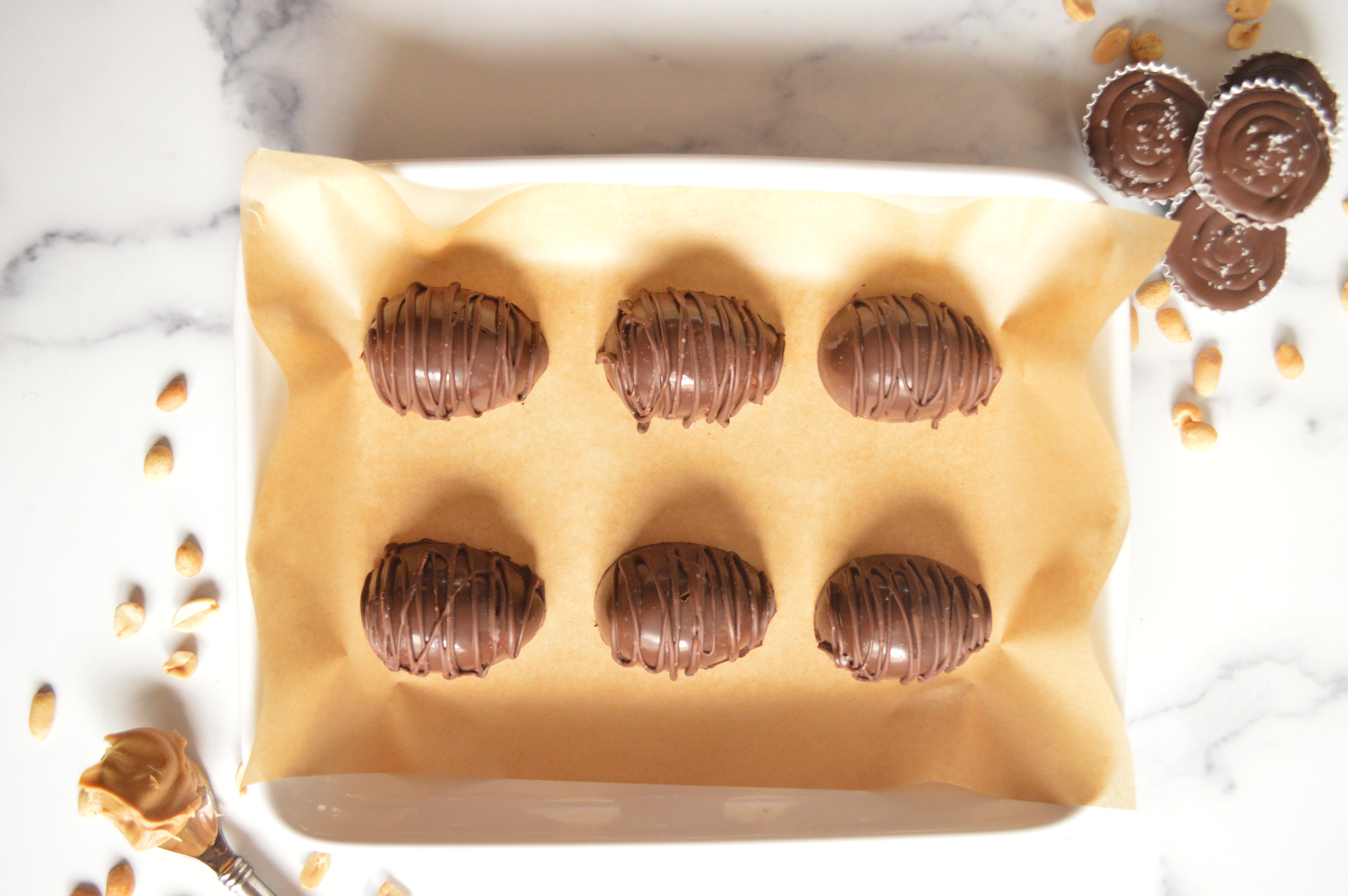 Peanut Butter Eggs on brown parchment paper on a white square platter, surrounded by peanuts, a spoonful of peanut butter and more peanut butter cups.