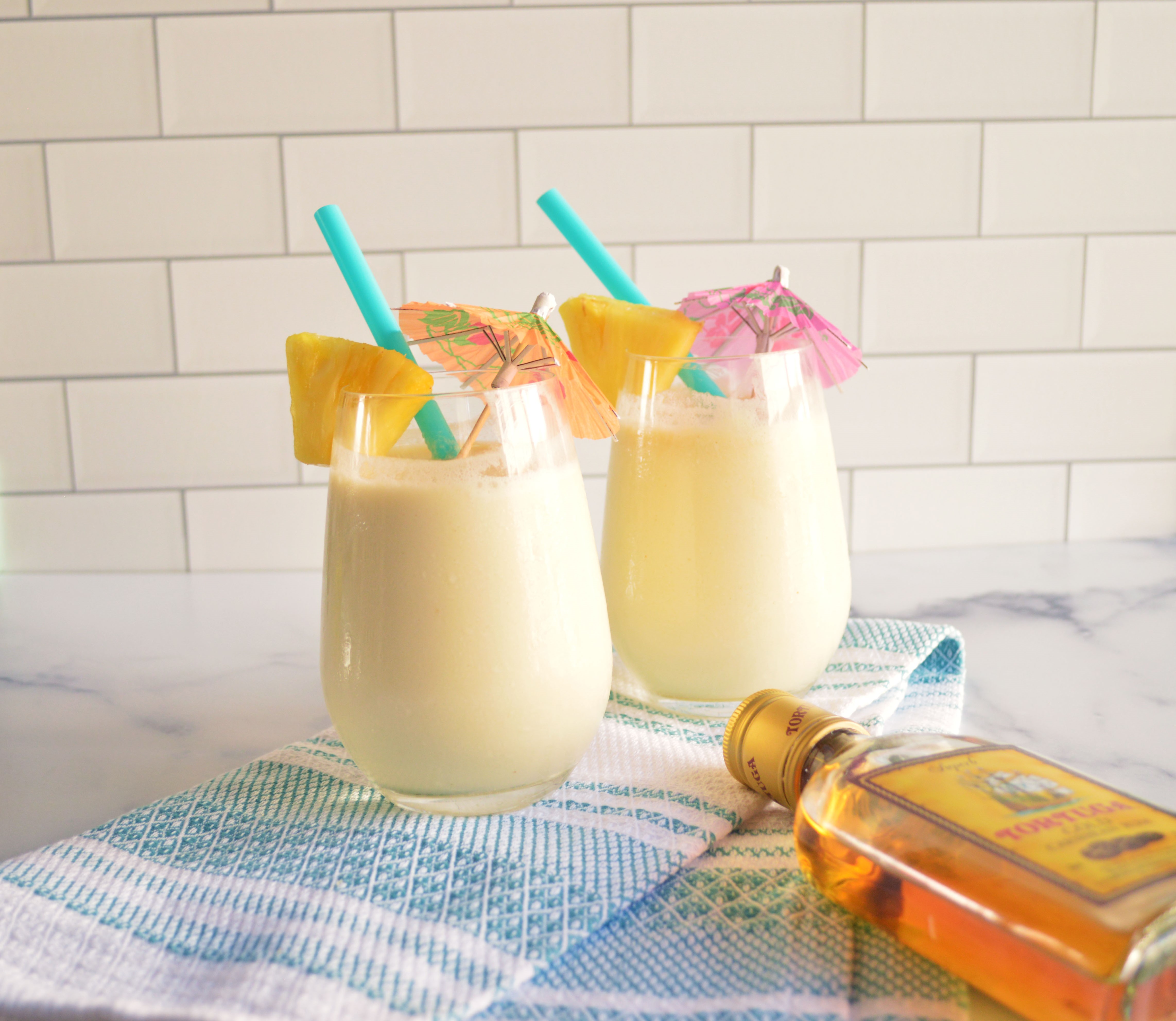 Two Piña Coladas sitting on a white and blue dish cloth like a towel at the beach with a bottle of Tortuga rum in the foreground 