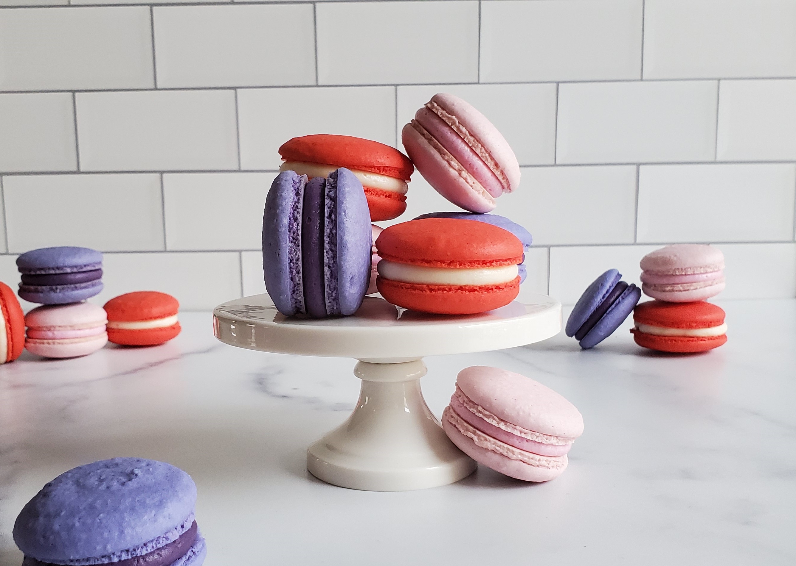 Purple, pink and red macarons, on a mini pedestal in front of a subway tile backsplash and more macarons in the background on a marble countertop.