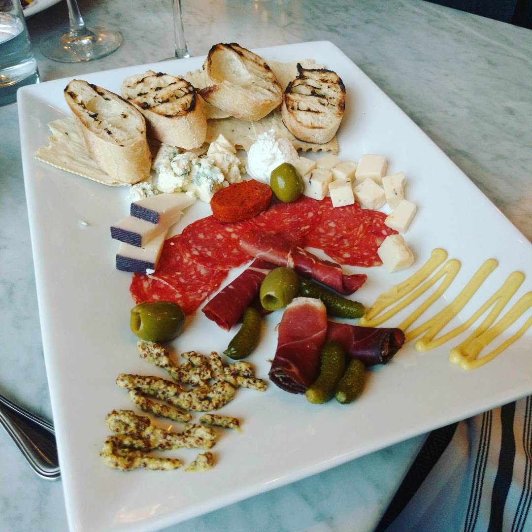 A charcuterie board with toasted bread, prosciutto, cheeses, pickles, olives, and mustard. On a square white plate and a white granite table top.