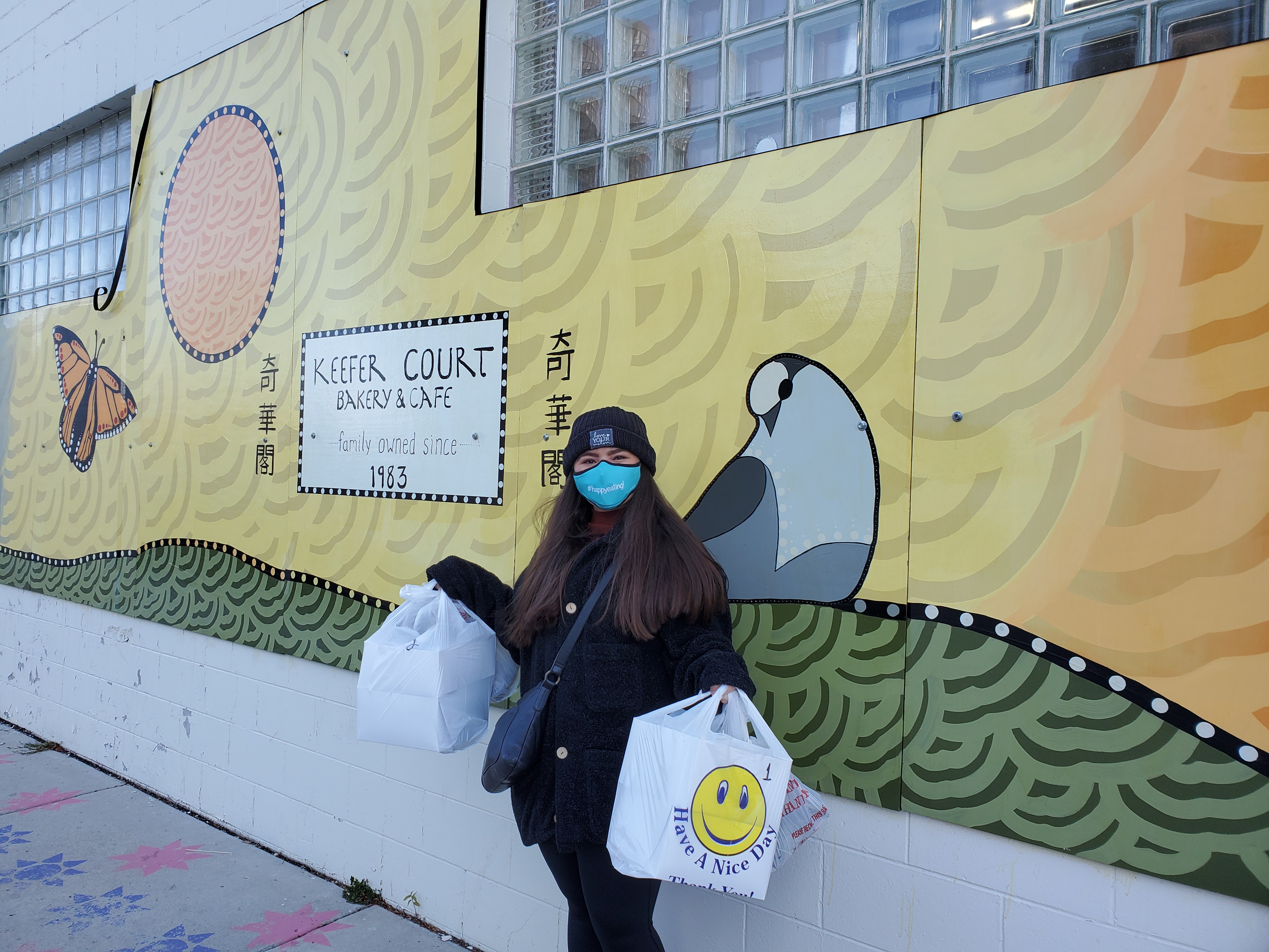 Me in front of the mural at Keefer Court holding huge bags of take out. The mural behind me reads "Keefer Court family owned since 1983".