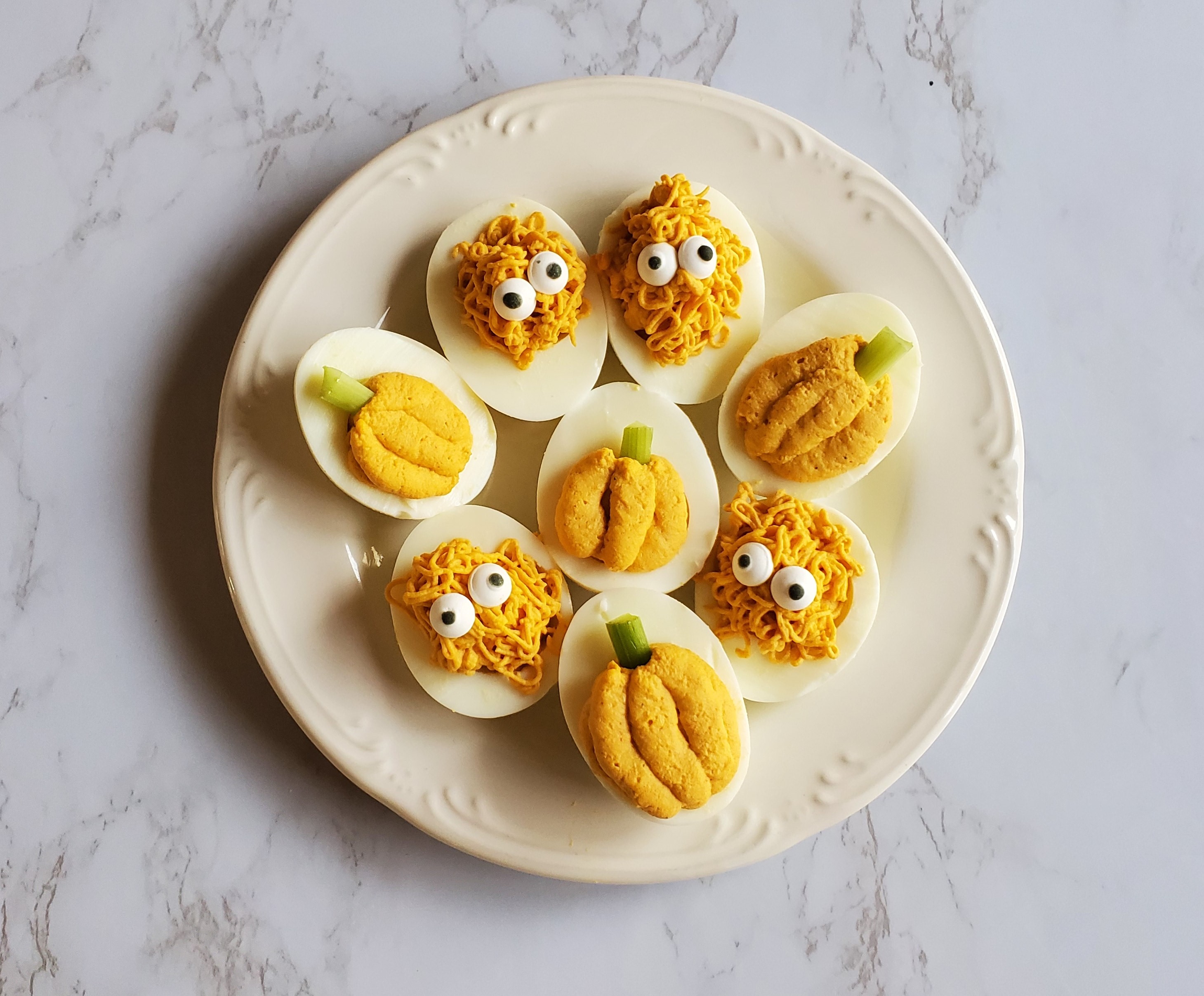 A white ceramic plate on a marble counter top filled with hard boiled eggs decorated as pumpkins and monsters using frosting piping tips..