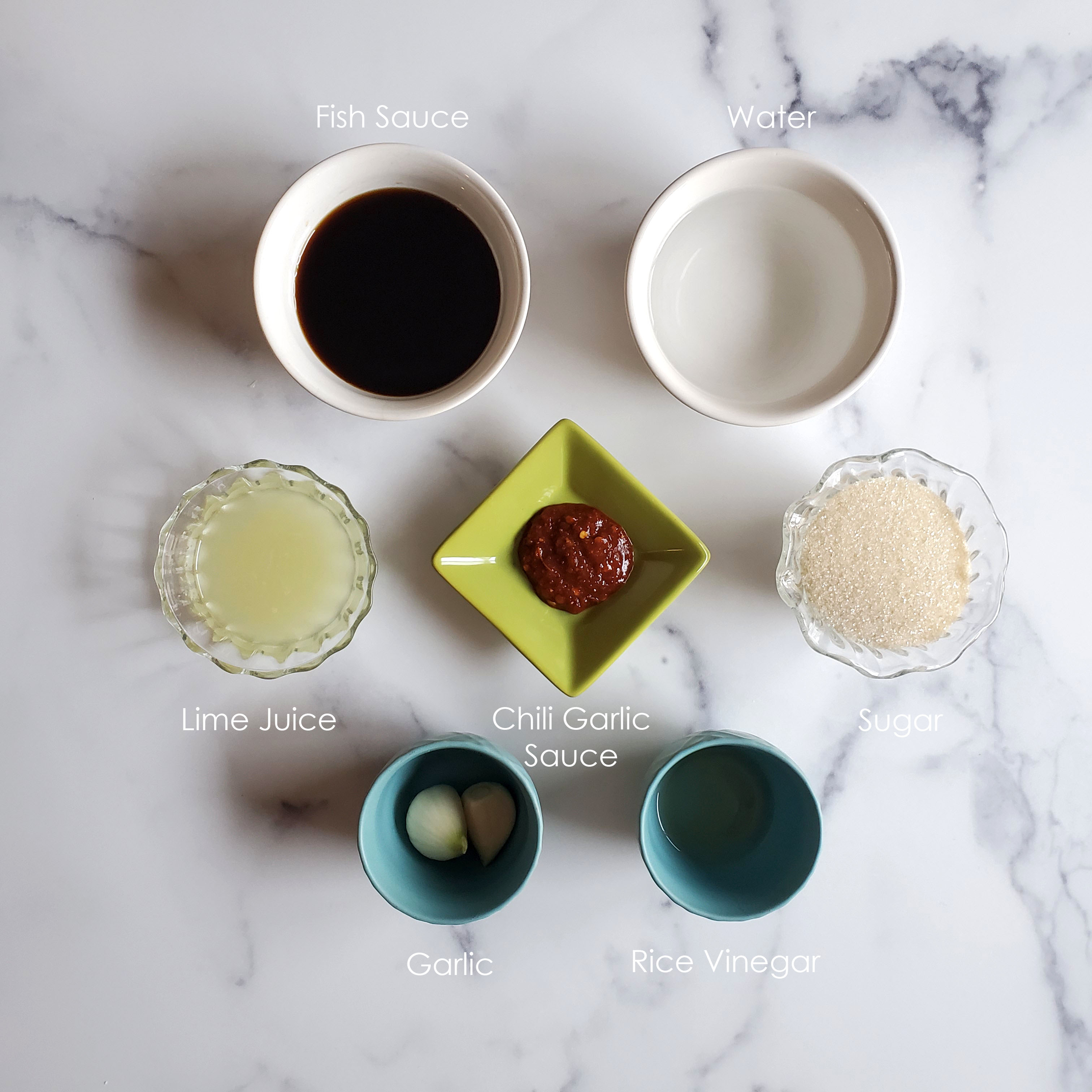 All of the ingredients are in bowls against a white marble background, fish sauce, water, lime juice, chili garlic sauce, sugar, garlic and rice vinegar.