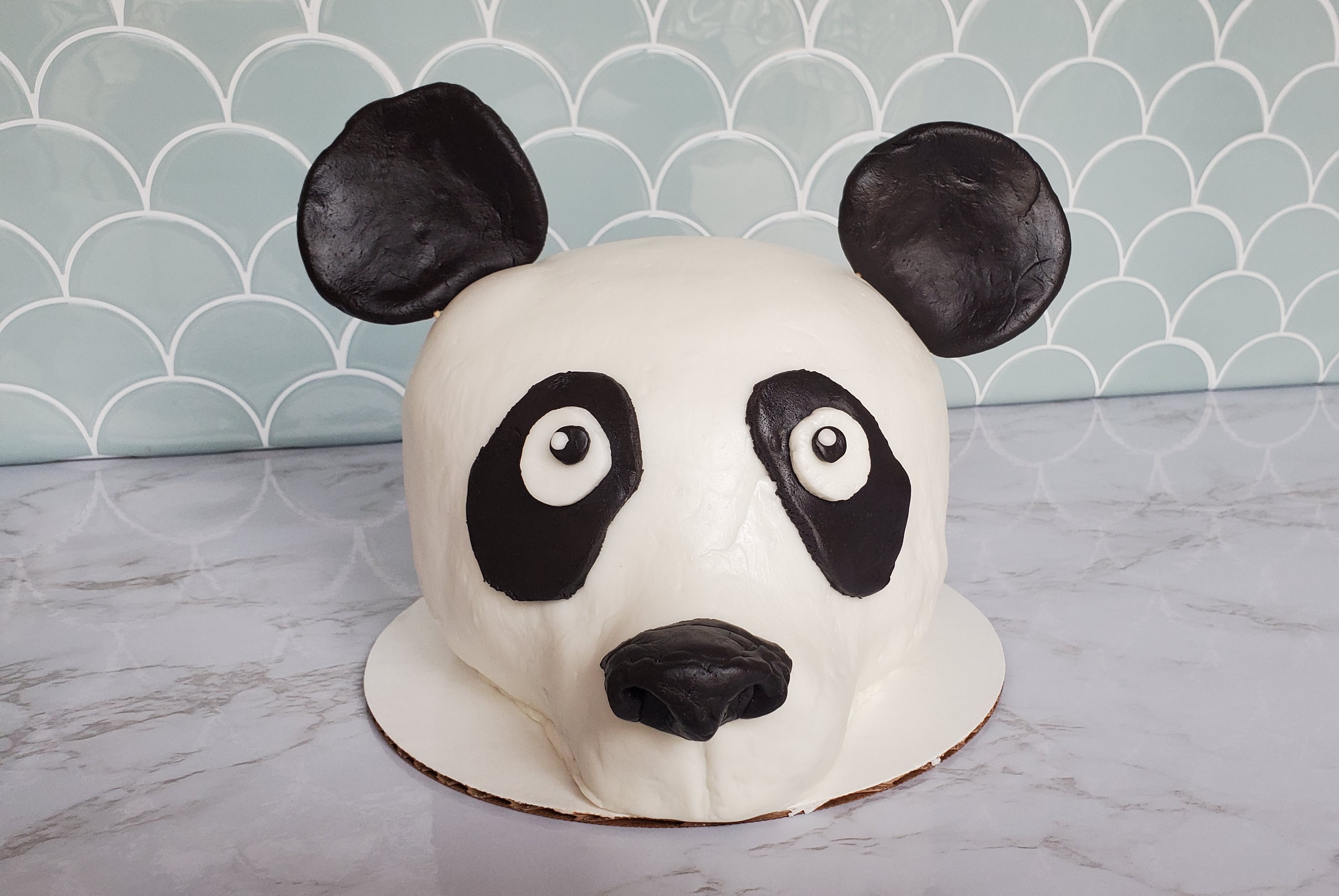 A cake that is decorated with fondant to look like the head of a panda. On a white marble countertop with a turquoise shell tile background.